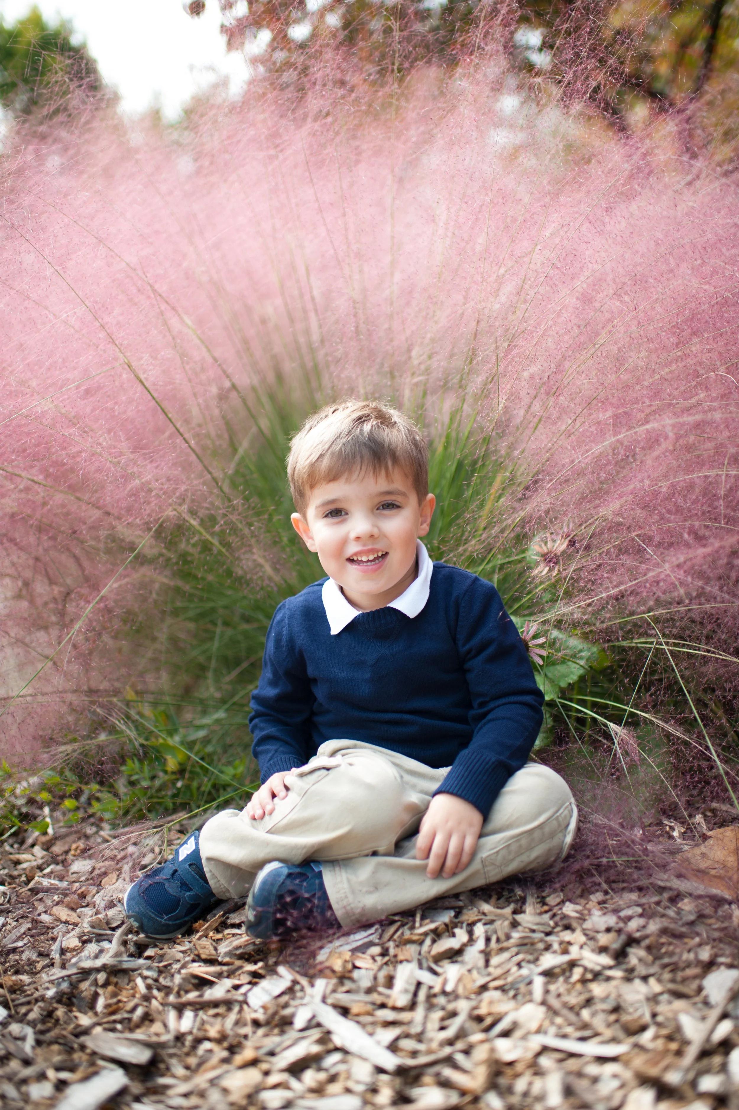 A young boy sitting on the ground outdoors on wood chips, smiling at the camera with pink ornamental grass behind him.