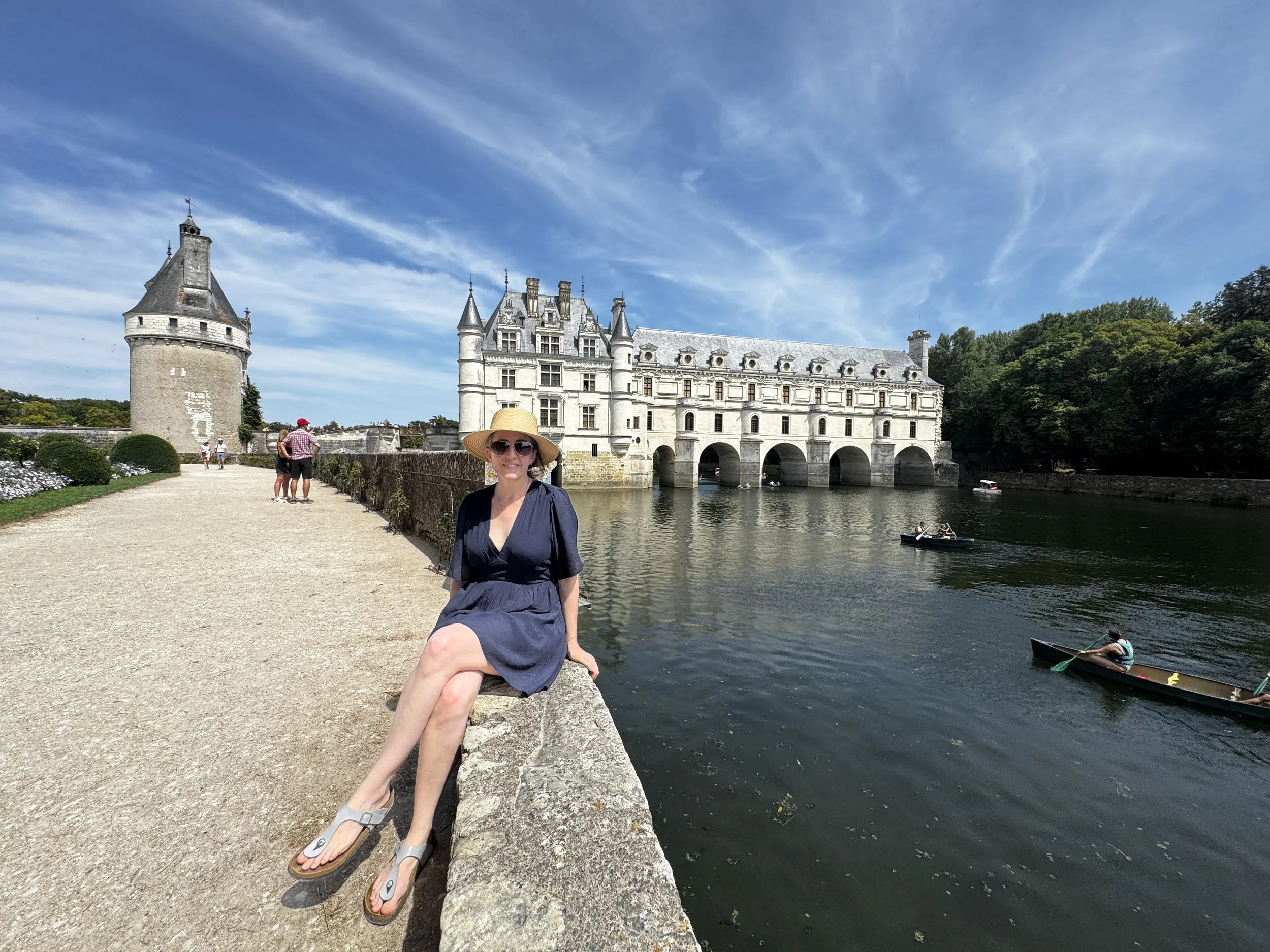 A woman sitting on a stone ledge near a river in front of a castle with multiple spires and turrets, surrounded by greenery and under a blue sky with wispy clouds, while others are walking and boating in the background.