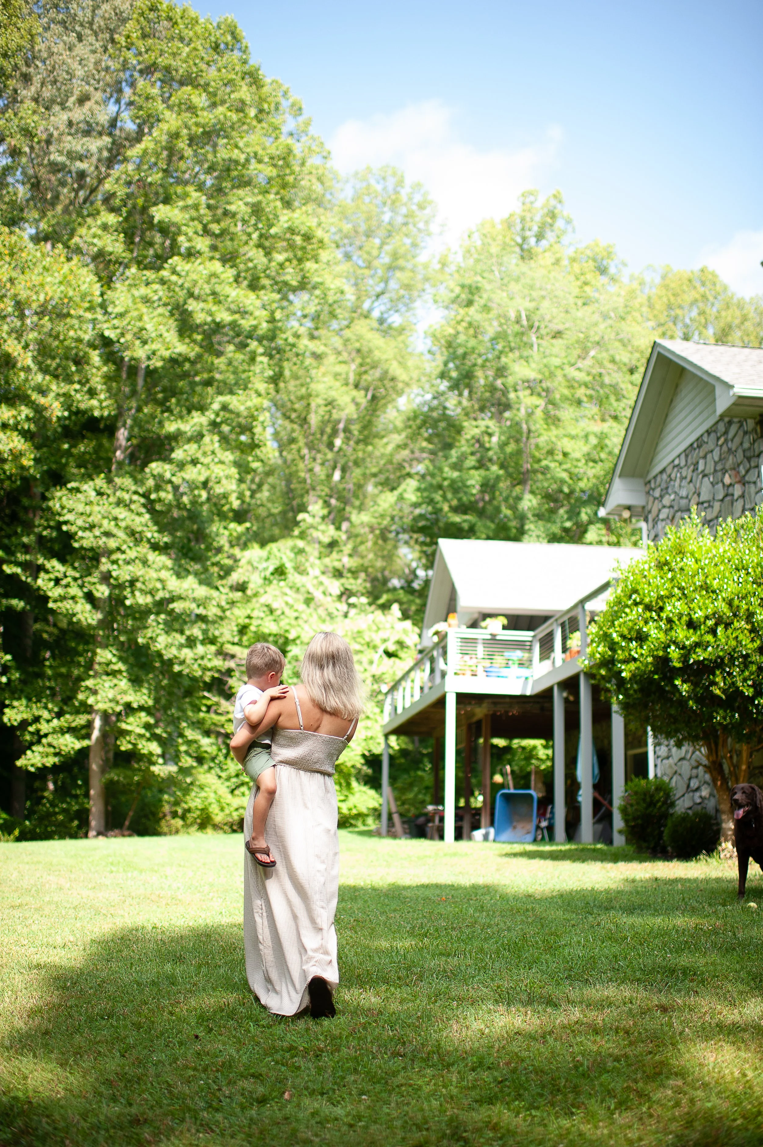 A woman with blonde hair holding a small boy in her arms on a lush green lawn with trees and a house in the background.