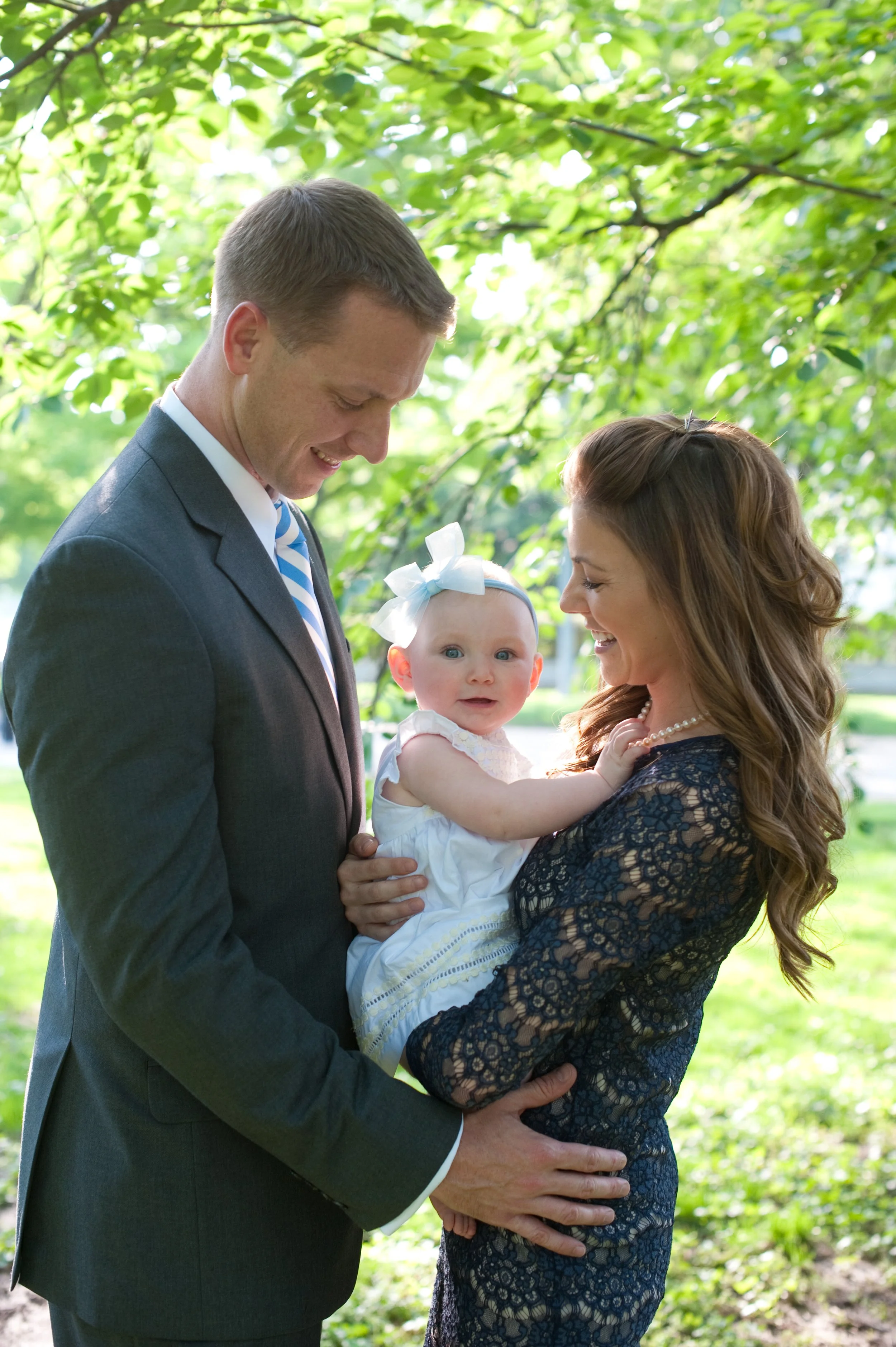 A young family outdoors, with a man in a suit, a woman in a blue lace dress, and a baby girl with a white bow, smiling and sharing a joyful moment under green trees.