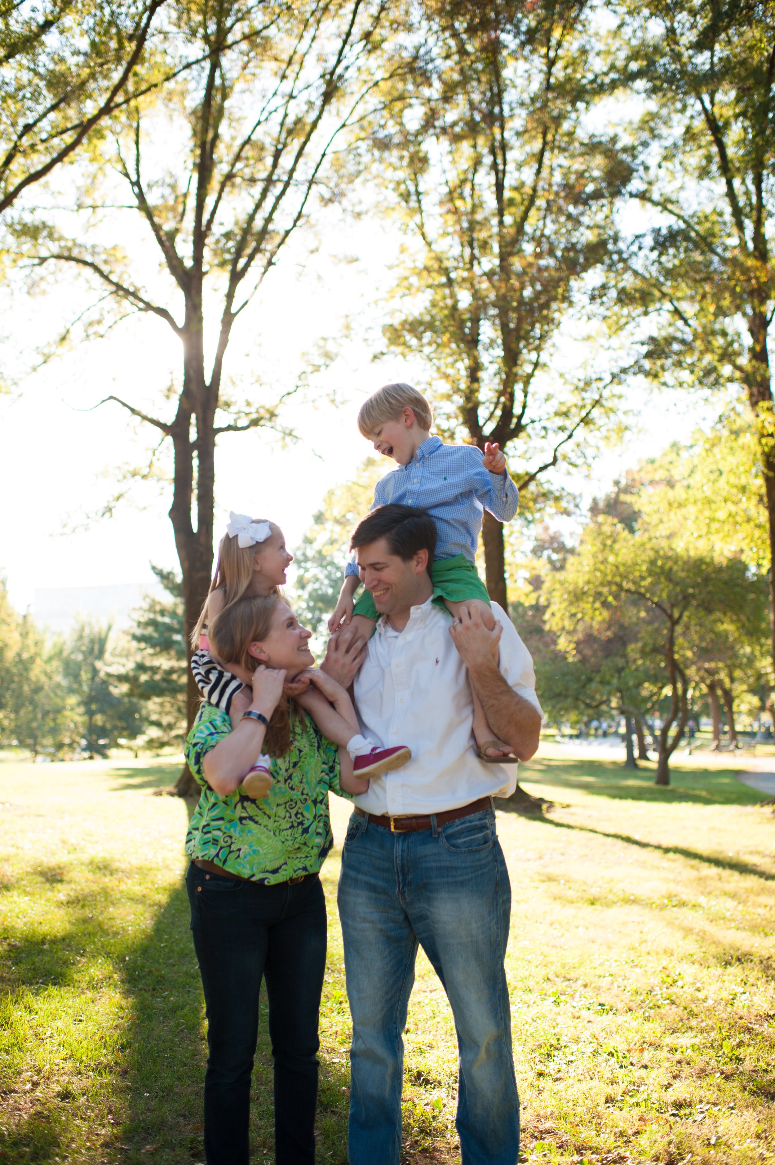 A family of four enjoying a day outdoors in a park with trees and sunlight, with a father carrying a young boy on his shoulders and a mother holding a young girl, all smiling and happy.