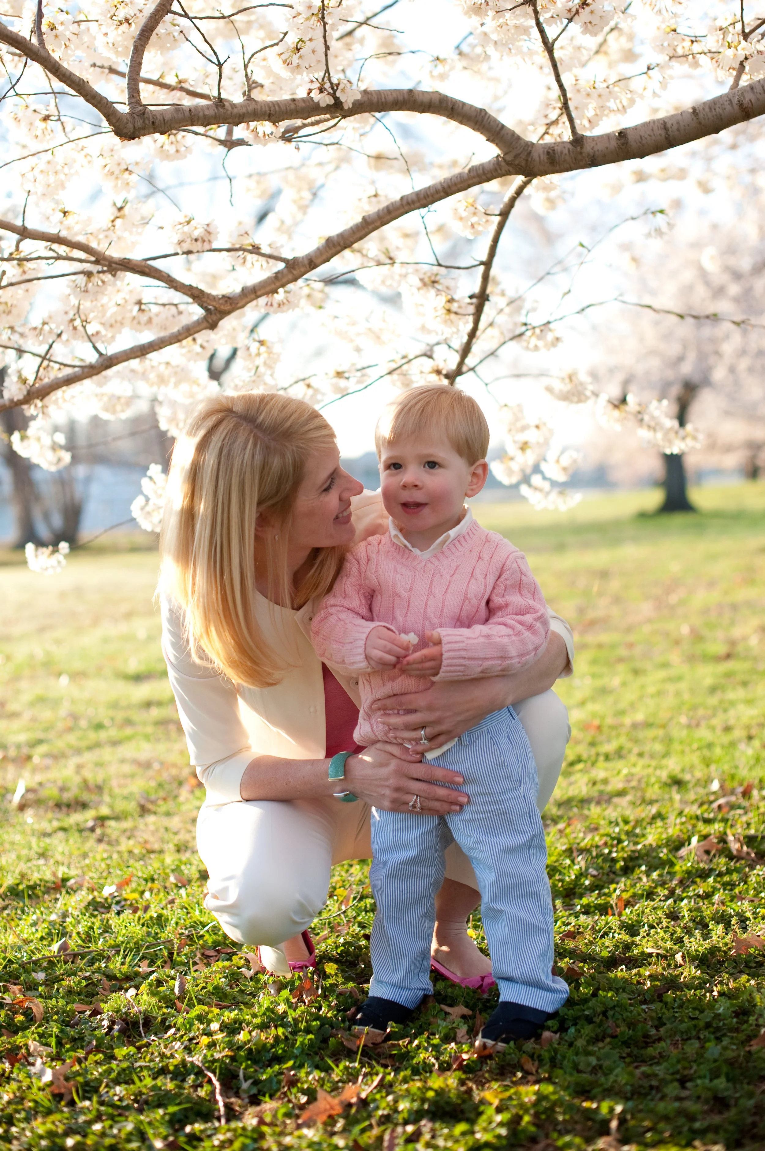 A woman and a young boy standing on grass under blooming cherry blossom trees in a park on a sunny day.