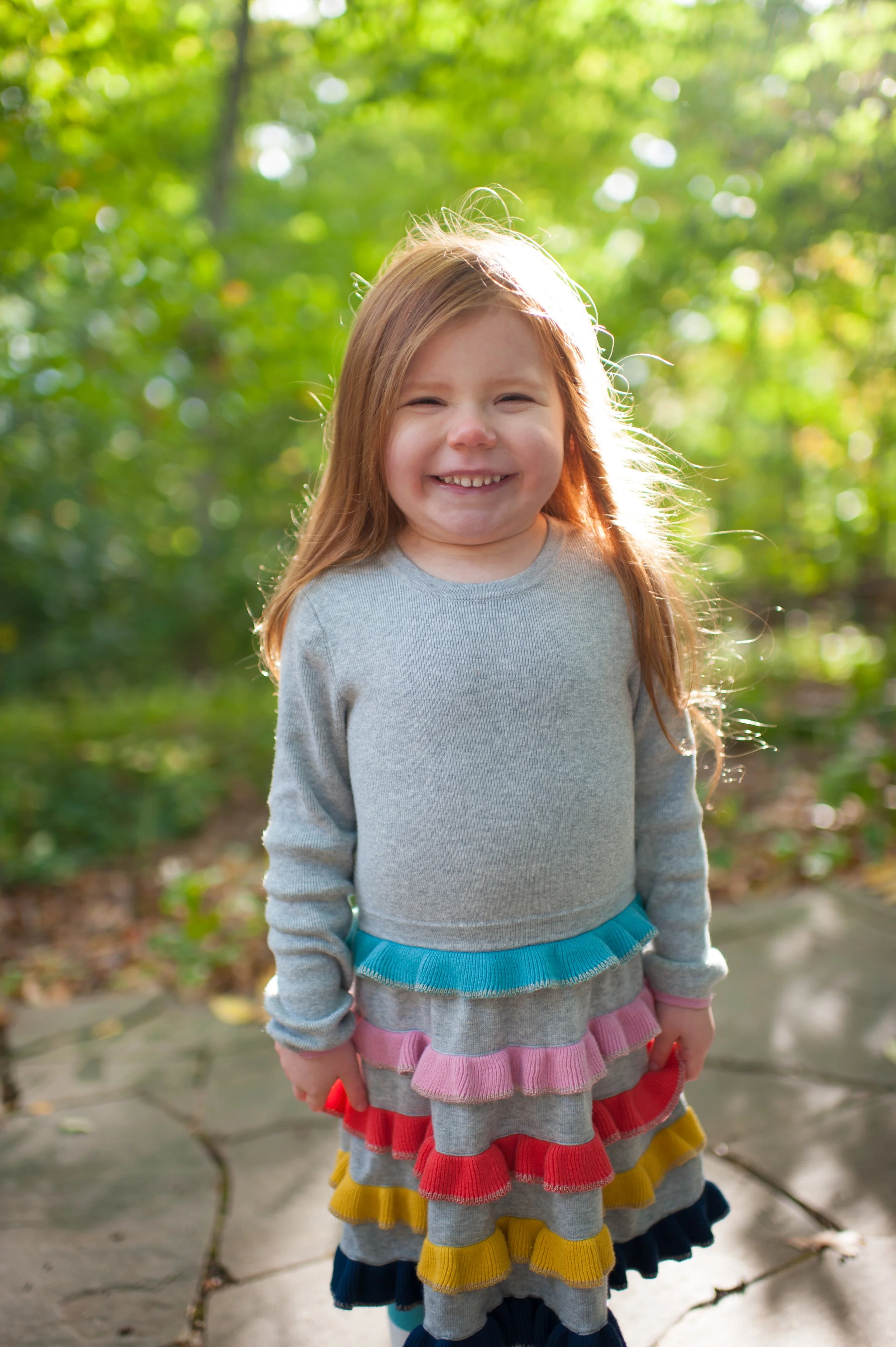 A smiling young girl with red hair standing outdoors on a sunny day, surrounded by green trees and bushes, wearing a grey long-sleeve shirt and a multicolored ruffled skirt.