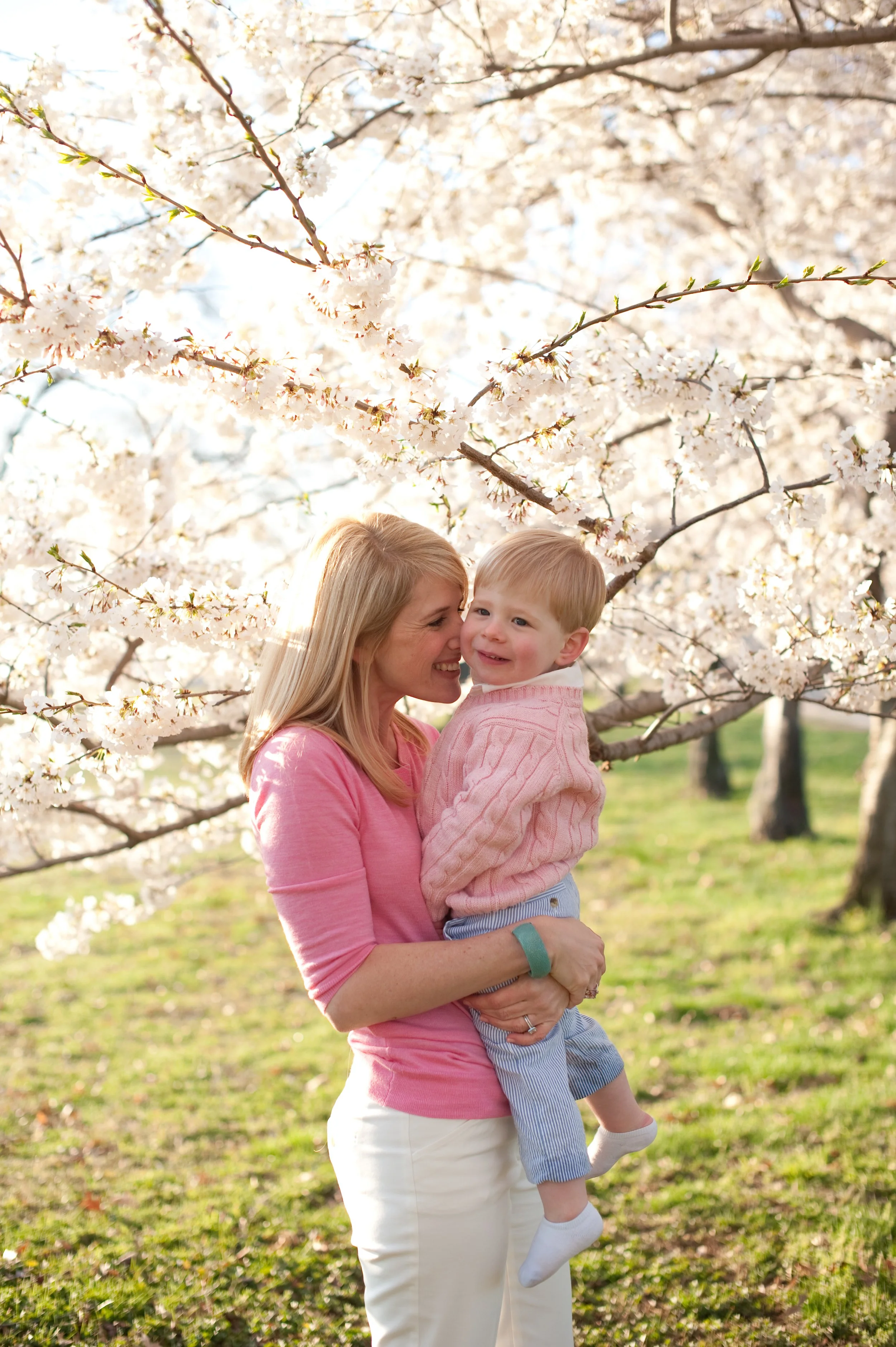 A smiling woman holding a young boy outdoors among blooming cherry blossom trees.