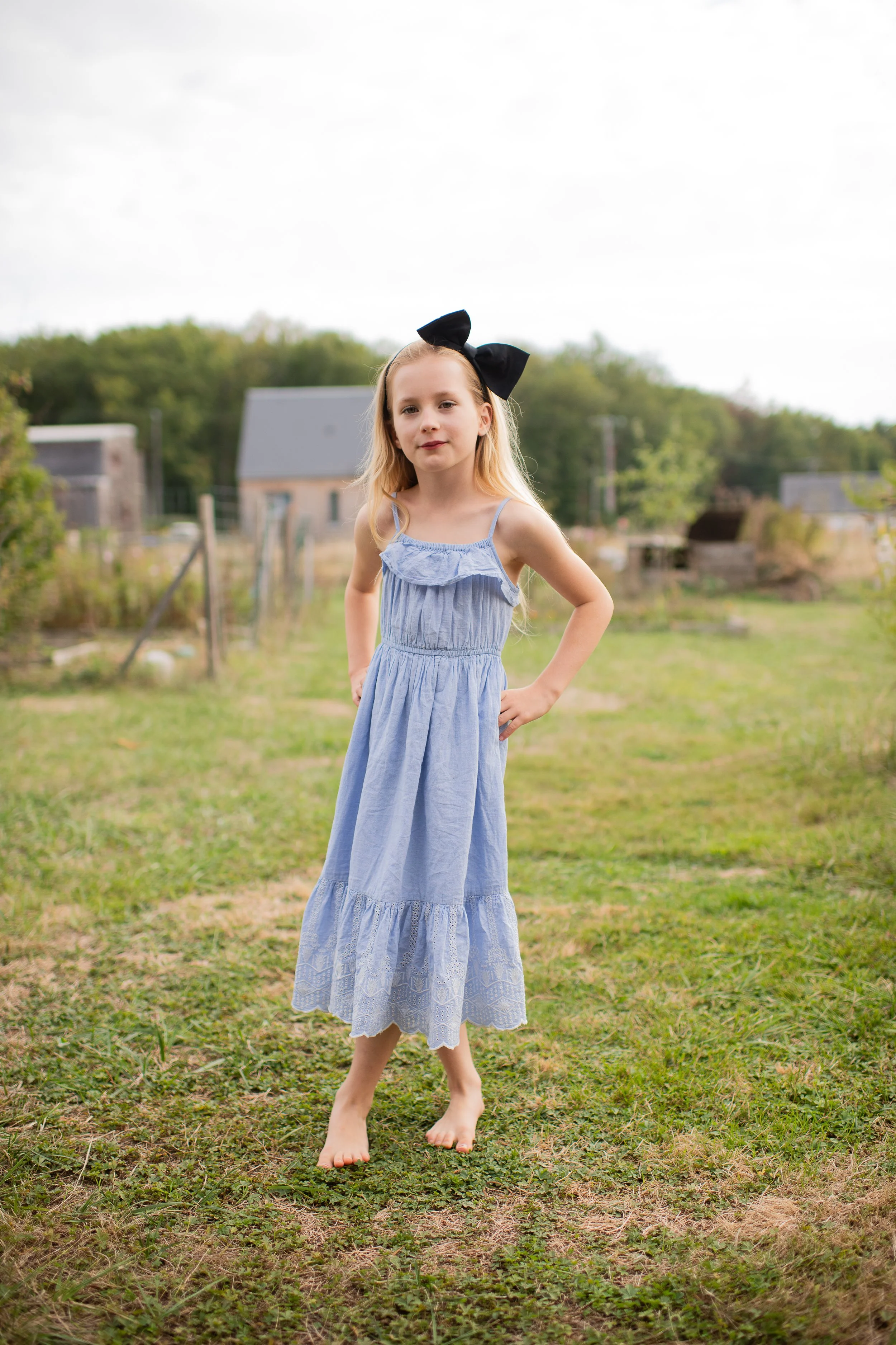A young girl with long blonde hair standing outdoors on grass, wearing a light blue sundress with straps and a ruffle detail, barefoot, with one hand on her hip and a large black bow in her hair, against a background of green trees and houses under a