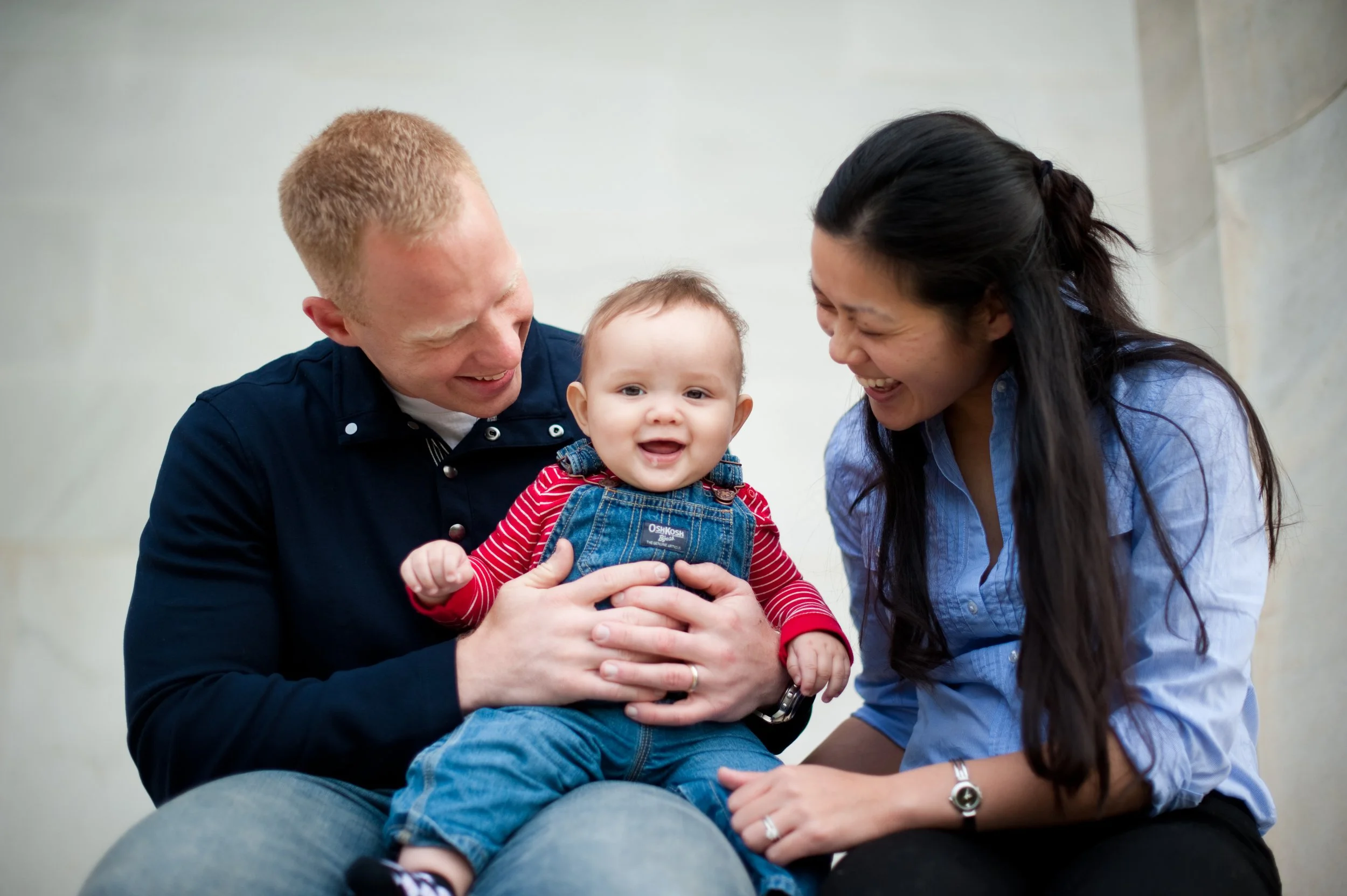 A happy family of three, a man, a woman, and a baby, sitting together and smiling indoors.
