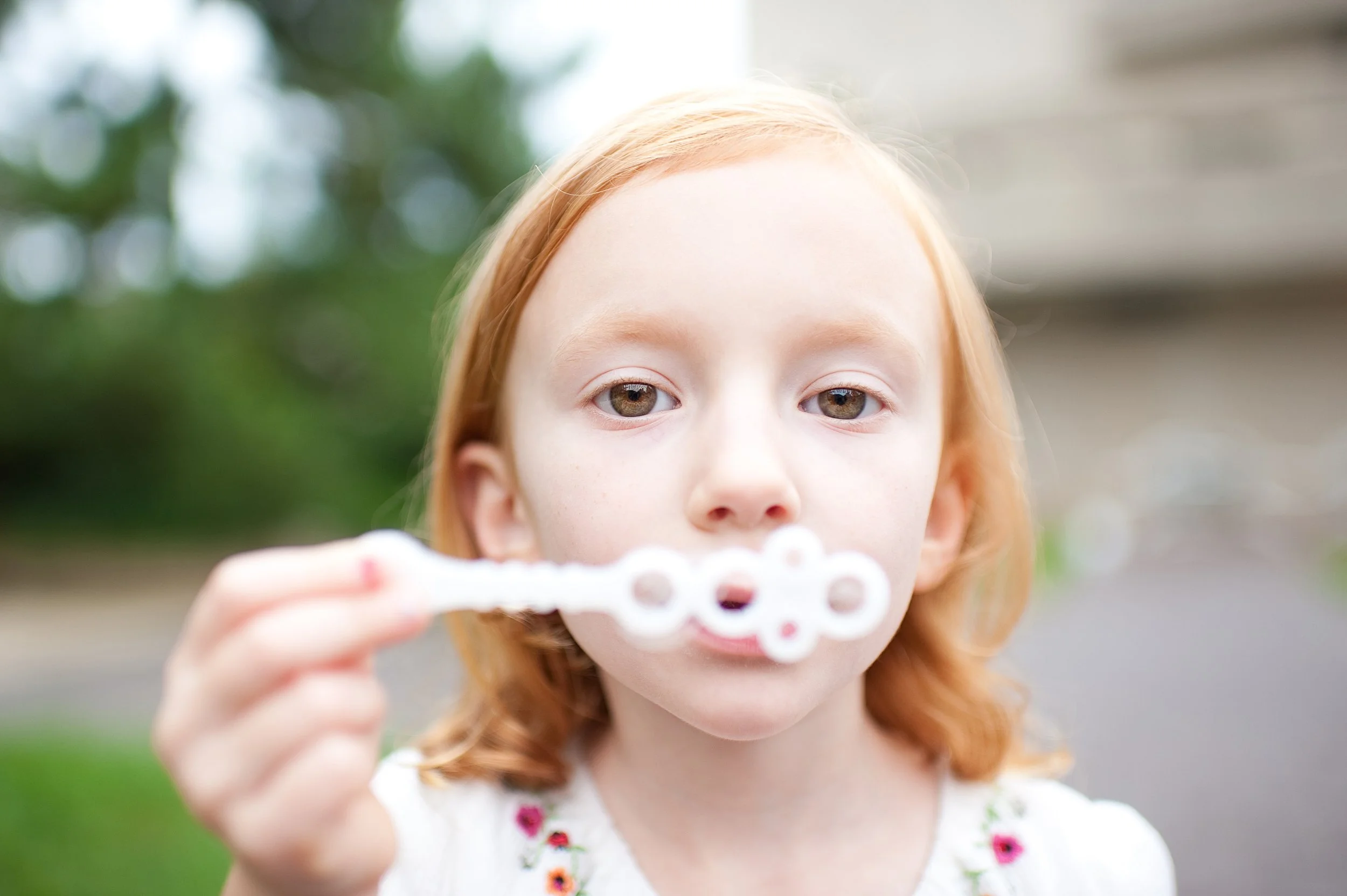 A young girl with red hair blowing bubbles outdoors, with a background of green trees and a building.