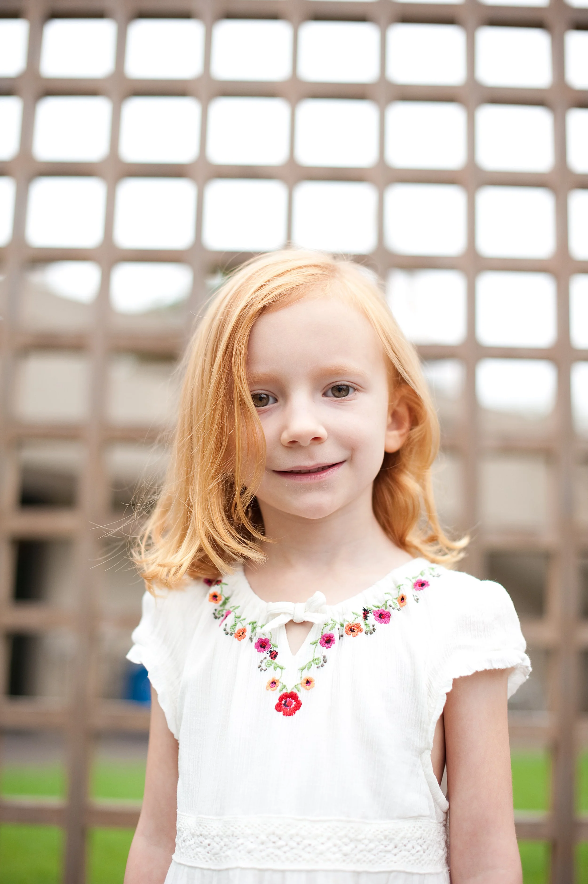 A young girl with red hair wearing a white dress with floral embroidery stands in front of a wooden trellis outdoor, smiling gently.