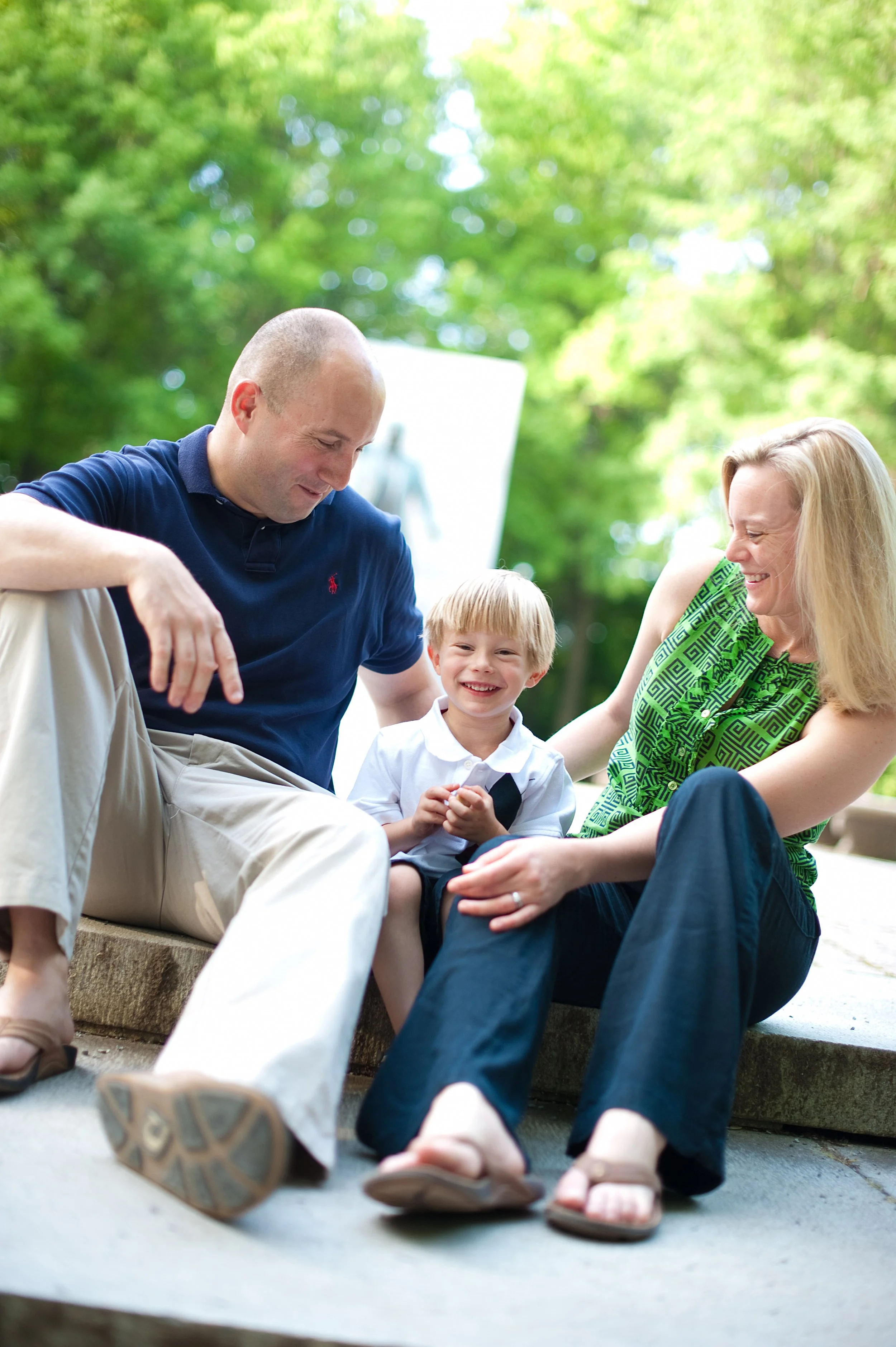 A family of three, with a man, woman, and young boy, sitting on a park bench outdoors, smiling and enjoying each other's company on a sunny day with green trees in the background.
