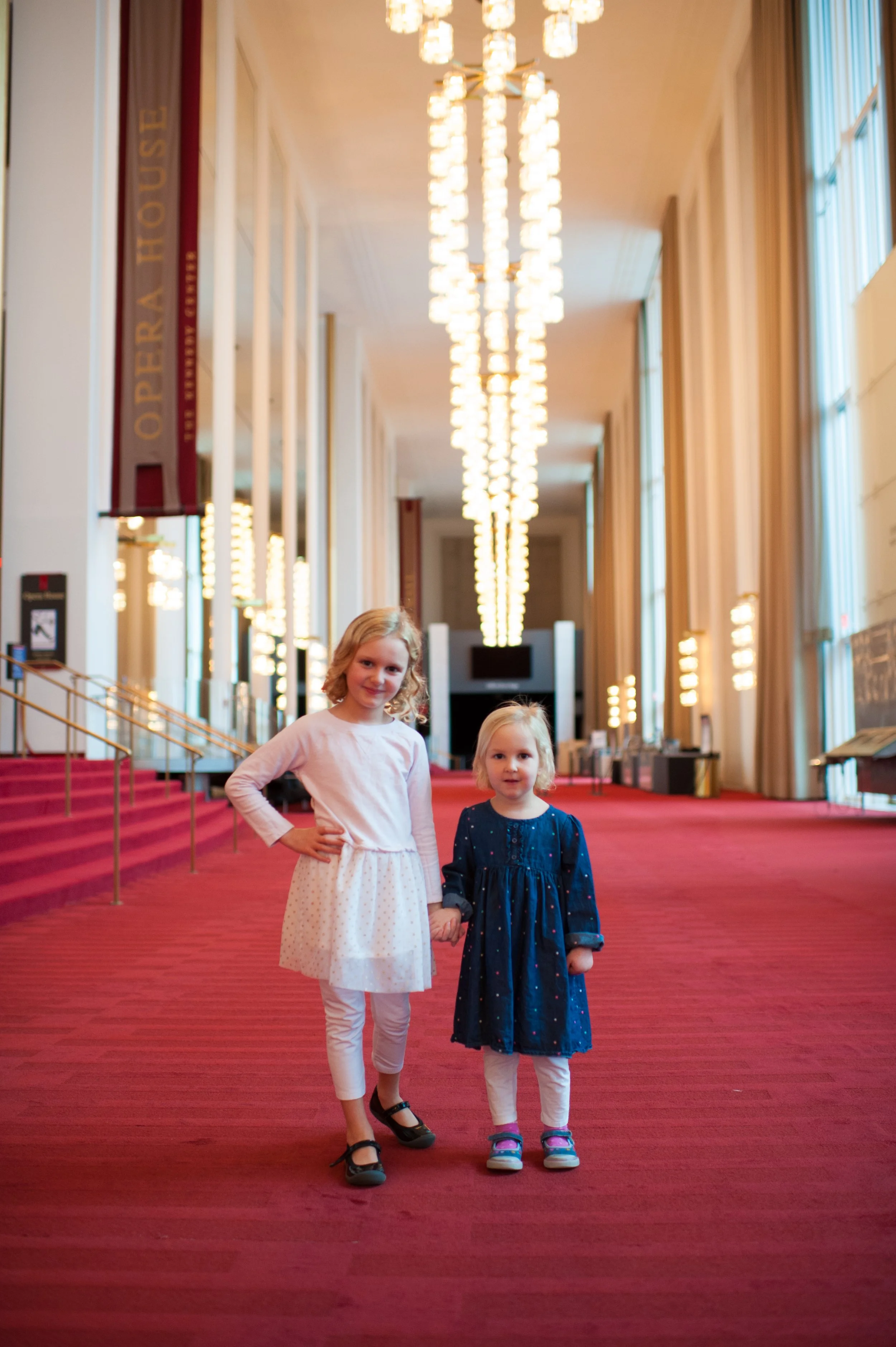 Two young girls holding hands and standing on a red carpeted hallway inside a building, with bright overhead lighting and large windows on the side.