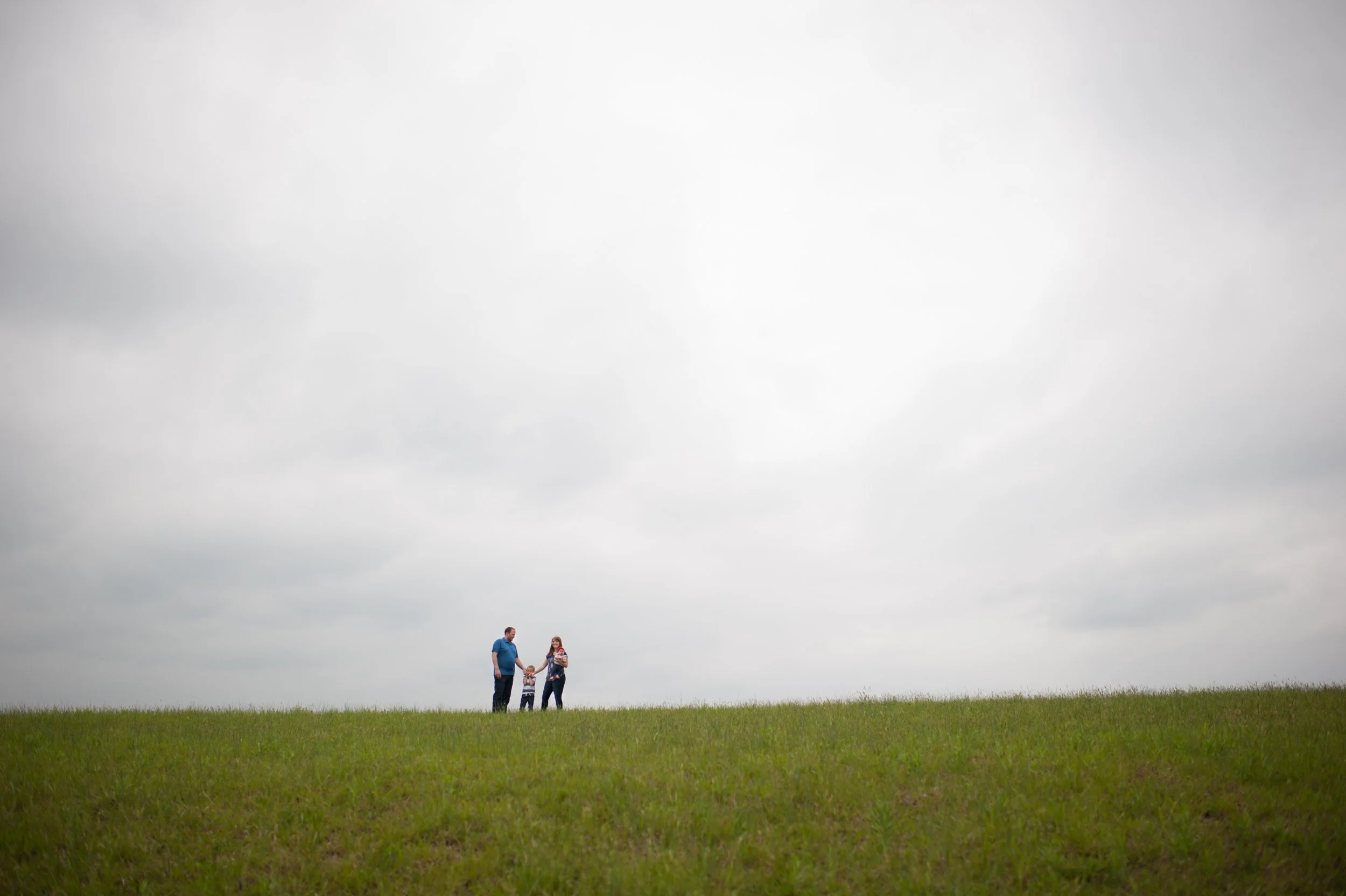 A family of four standing on a grassy hill against a cloudy sky, holding hands.