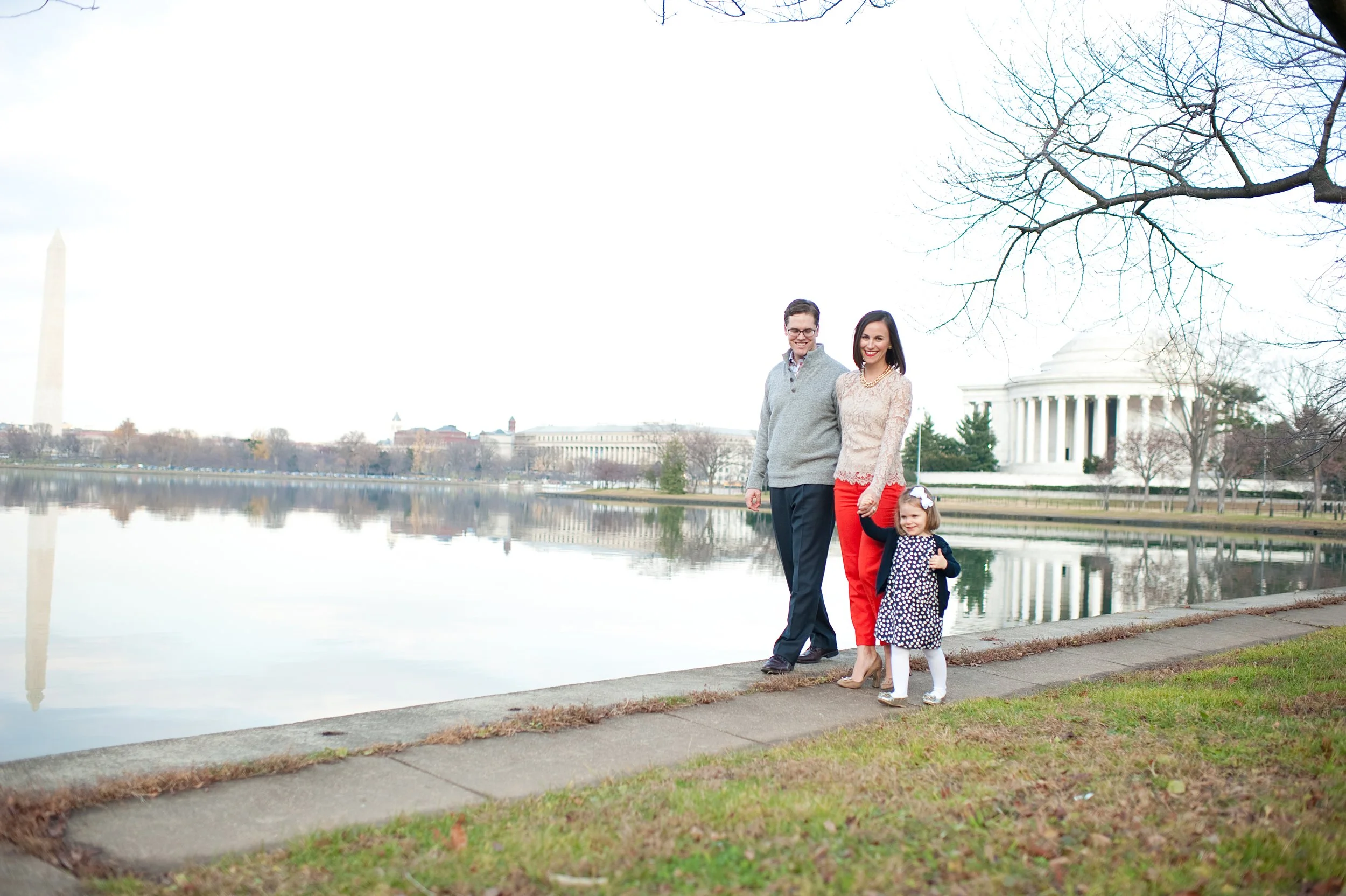 A family of three standing on a sidewalk near a body of water, with the Washington Monument and the Jefferson Memorial visible in the background, during daytime.