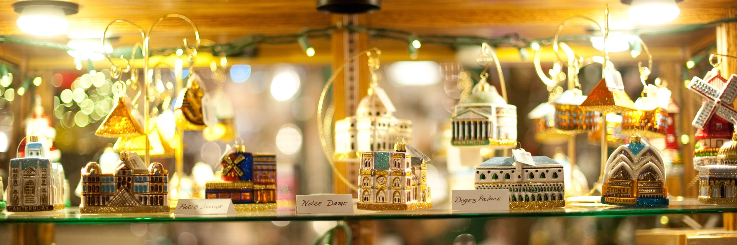 Collection of miniature souvenir buildings and landmarks including Canterbury, Paris Louvre, Notre Dame, Doge's Palace, and Sydney Opera House, displayed on a glass shelf with blurred festive lights in the background.