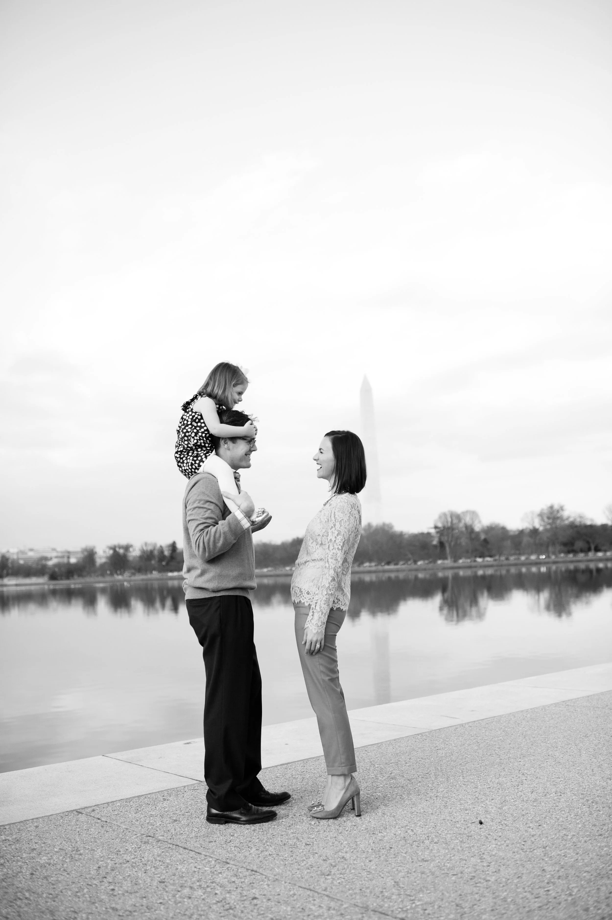 A black and white photo of a family by a lake, with the Washington Monument visible in the background. A man is holding a young girl on his shoulders, and a woman is standing facing them, smiling.