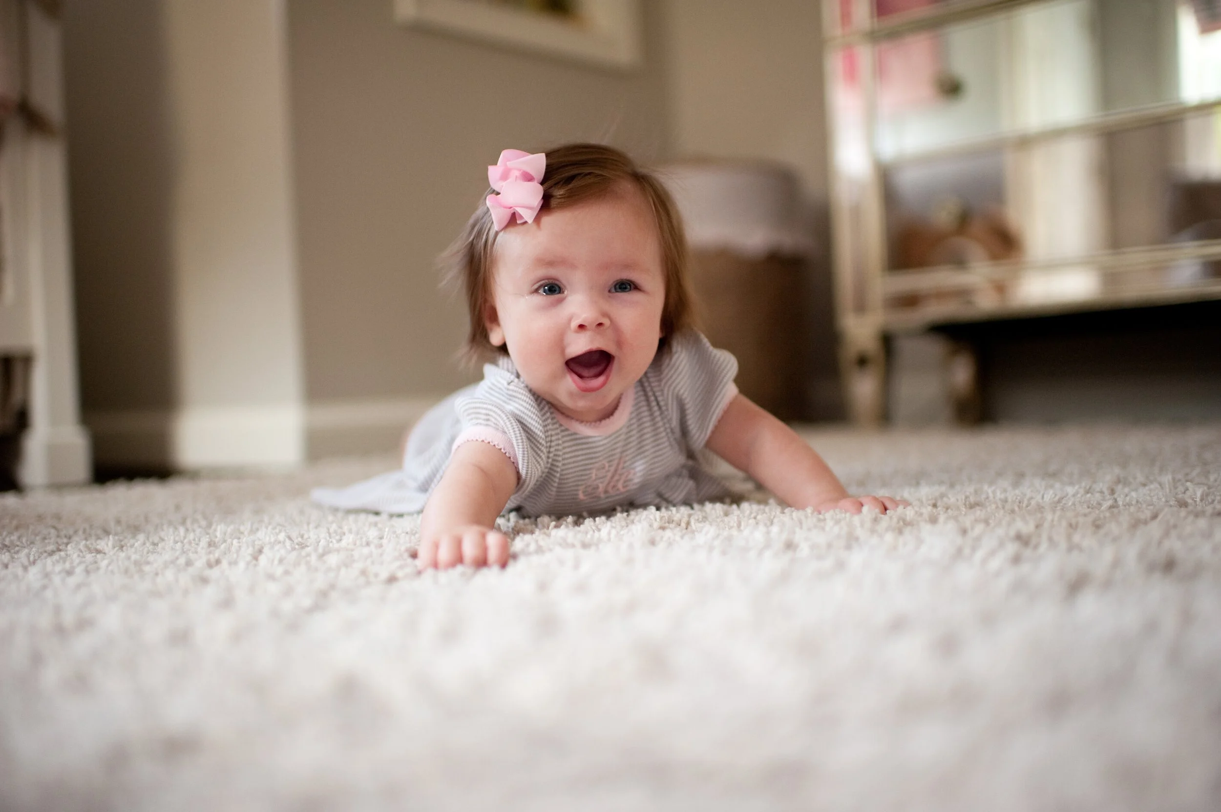 A baby girl with a pink bow in her hair crawling on a carpeted floor and smiling.