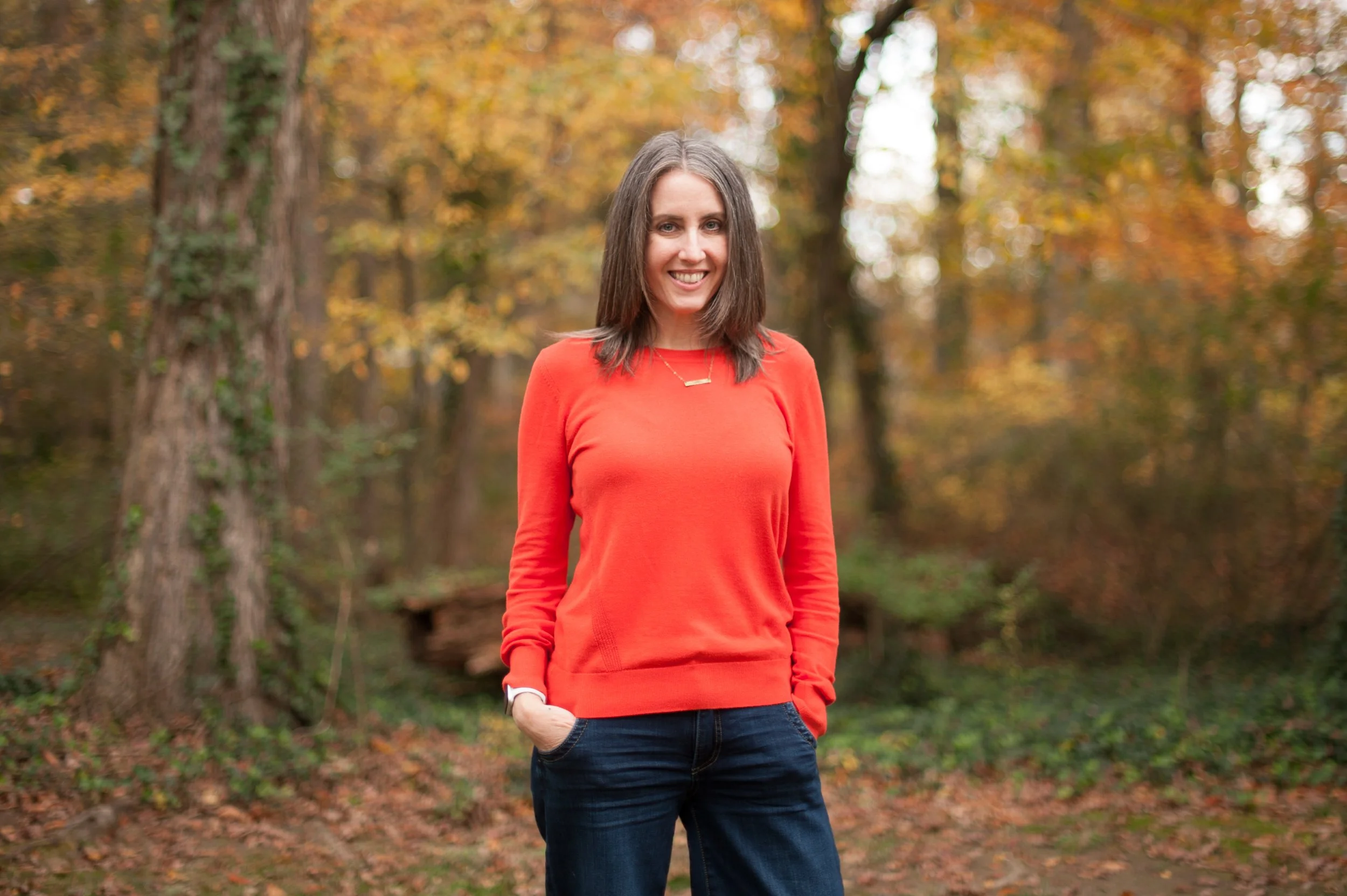 A woman standing outdoors in a forest with autumn foliage, wearing a red sweater and dark jeans, smiling with her hands in her pockets.