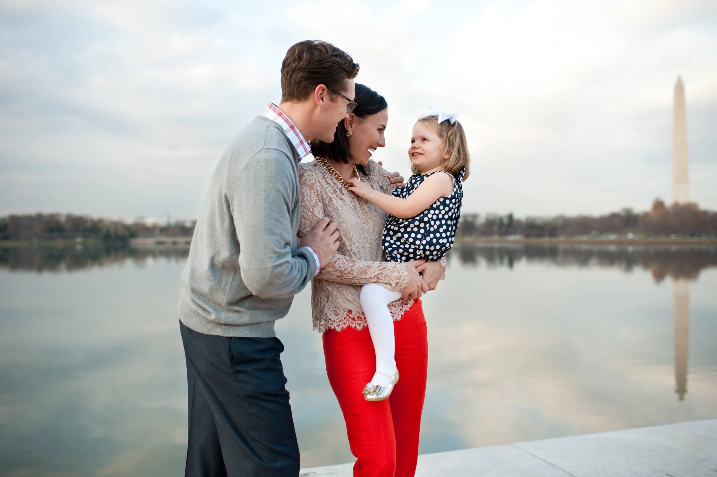 A family of three standing by the water, with the Washington Monument in the background. The mother holds a young girl, while the father looks at them smiling.