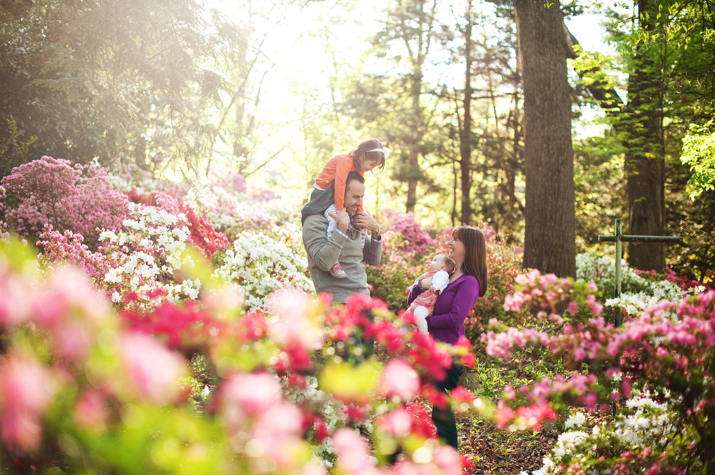 A family of four enjoying a walk in a blooming garden with pink and white flowers, sunlight filtering through trees, and a father giving his daughter a piggyback ride while the mother holds a baby.