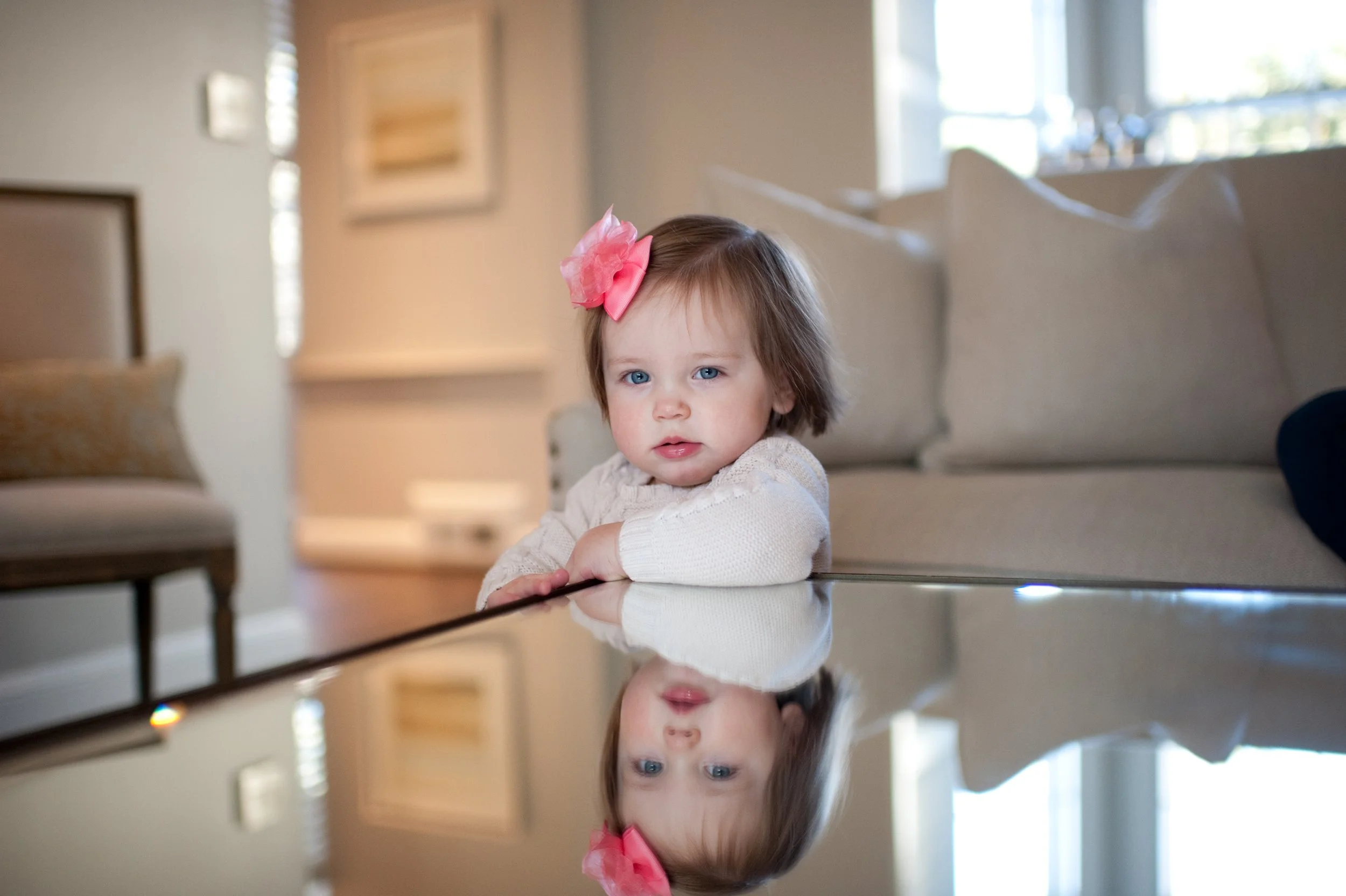 A young girl with blue eyes and brown hair, wearing a white sweater and a pink bow, rests her arms on a reflective surface in a living room with beige furniture and natural light.