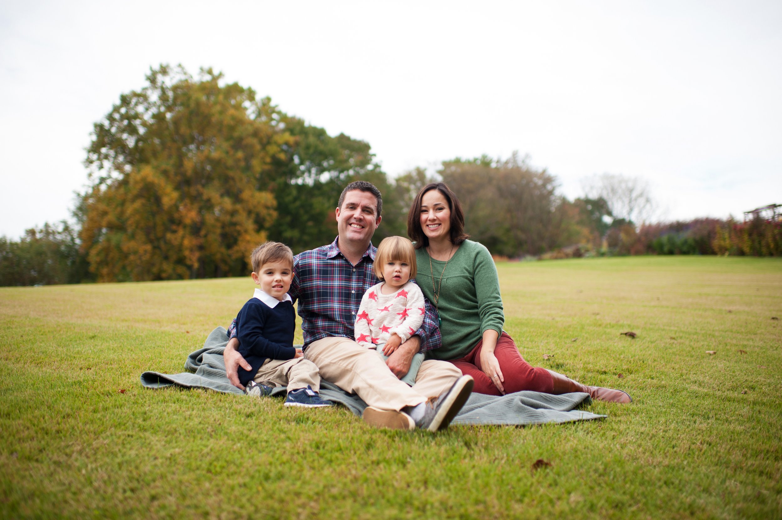 A family of four sitting on a blanket in a park with green grass and trees with fall foliage, smiling at the camera.