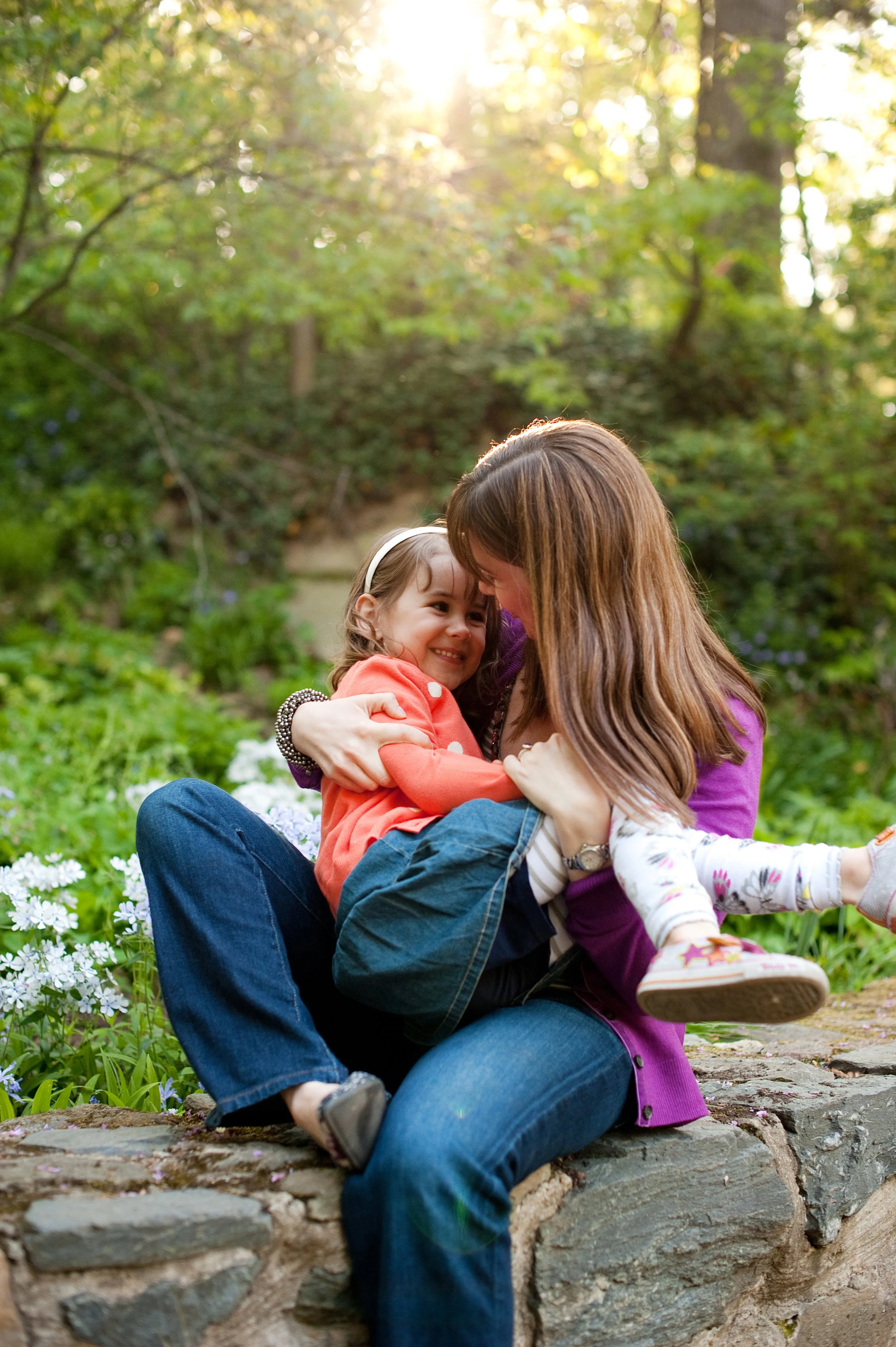 A woman and a young girl sharing an embrace outdoors in a park with greenery and blooming flowers, sun shining through trees.