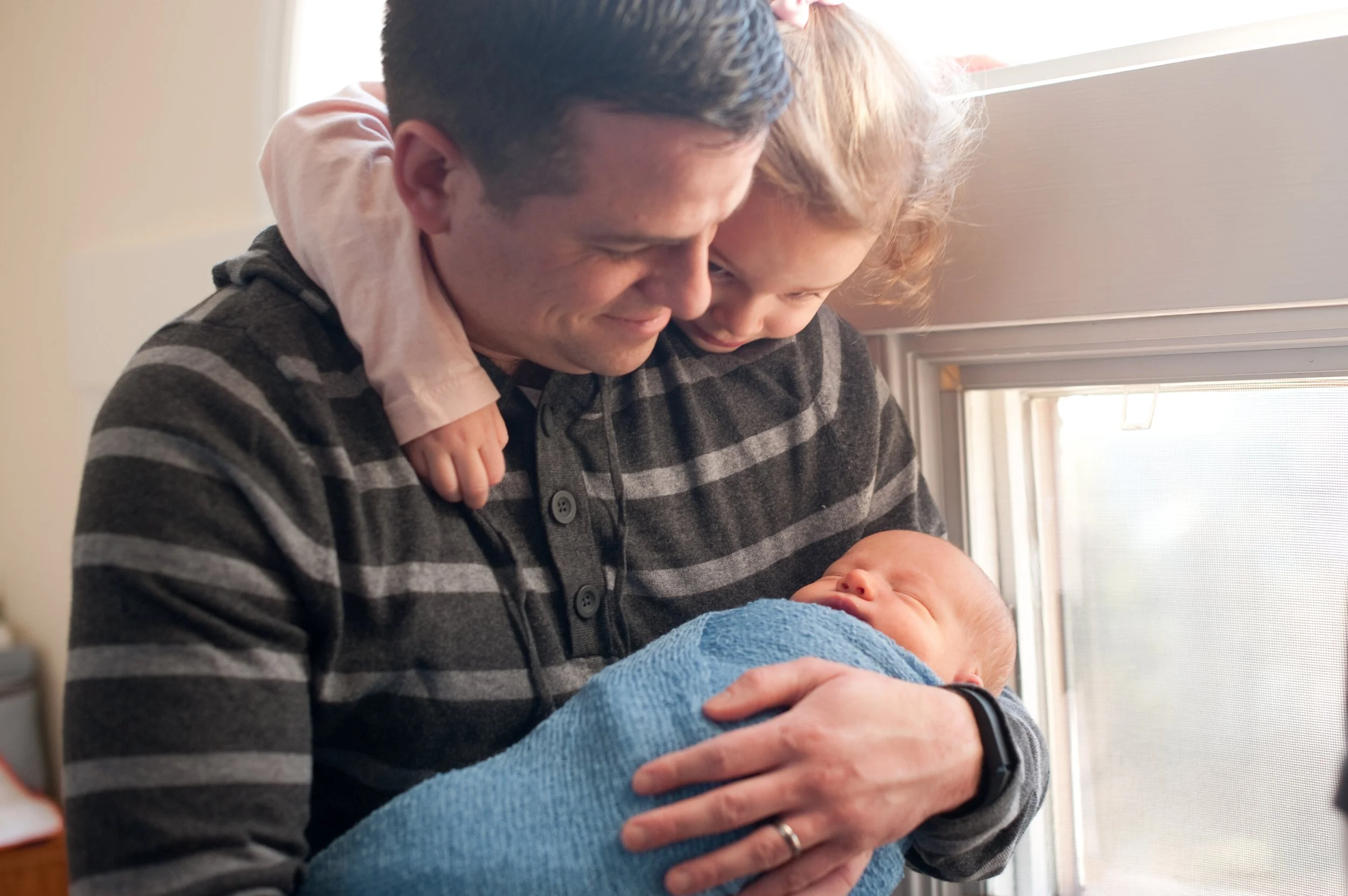 A man holding a newborn baby wrapped in a blue blanket, with an older child hugging him, in front of a window.