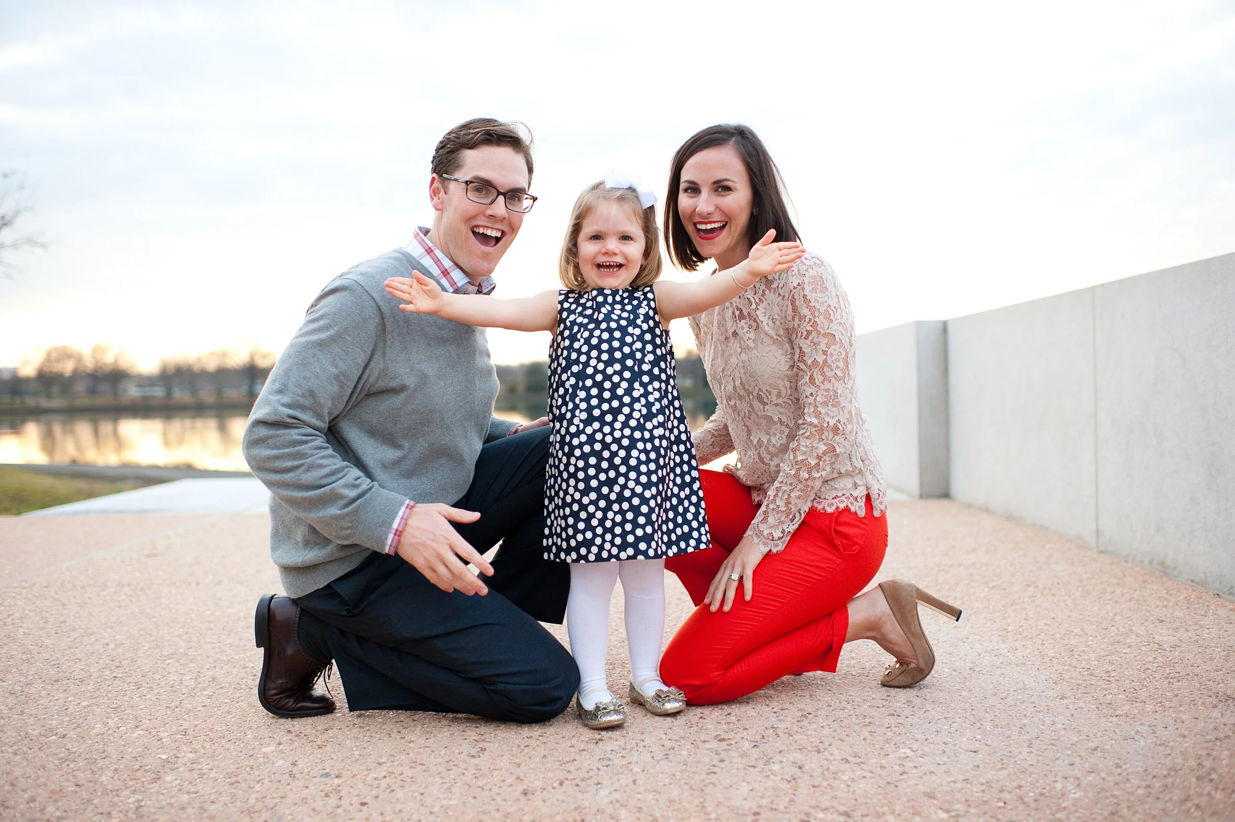 A happy family with a man, woman, and little girl outdoors near a body of water at sunset, smiling and posing together.