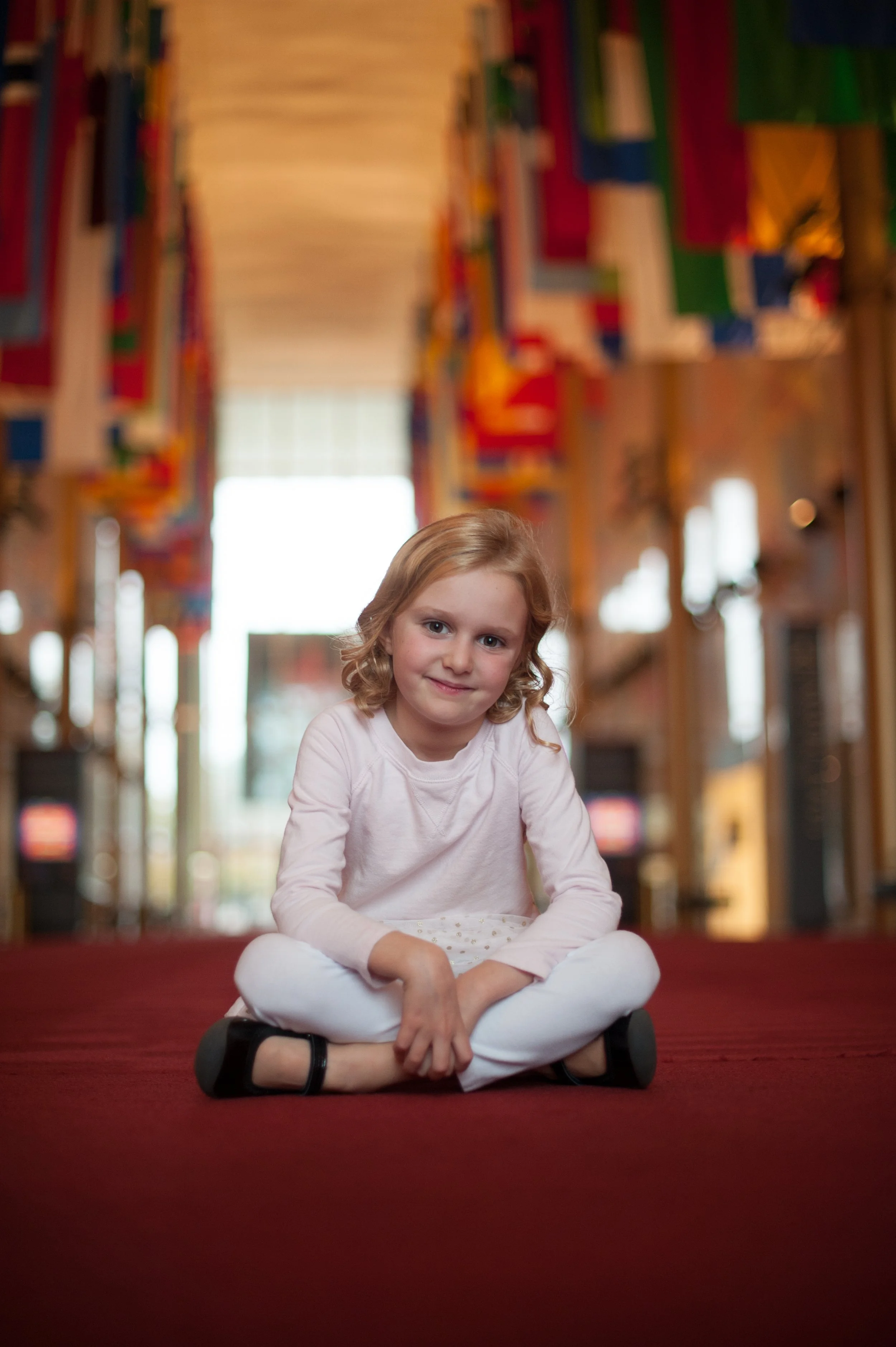 A young girl with blonde curly hair sitting cross-legged on a red carpet in a colorful indoor setting with abstract artwork or decorations hanging on the ceiling.