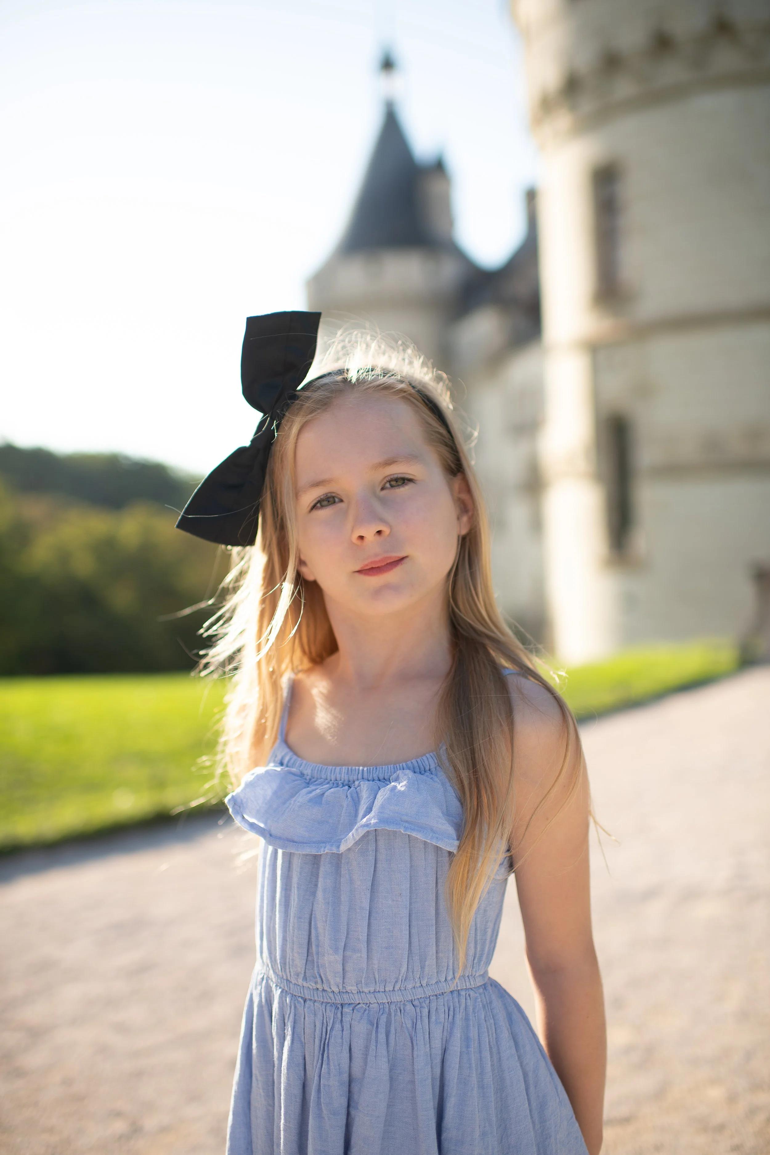 Young girl with long blonde hair wearing a light blue dress and a large black bow on her head standing outdoors near a historic castle in the background.