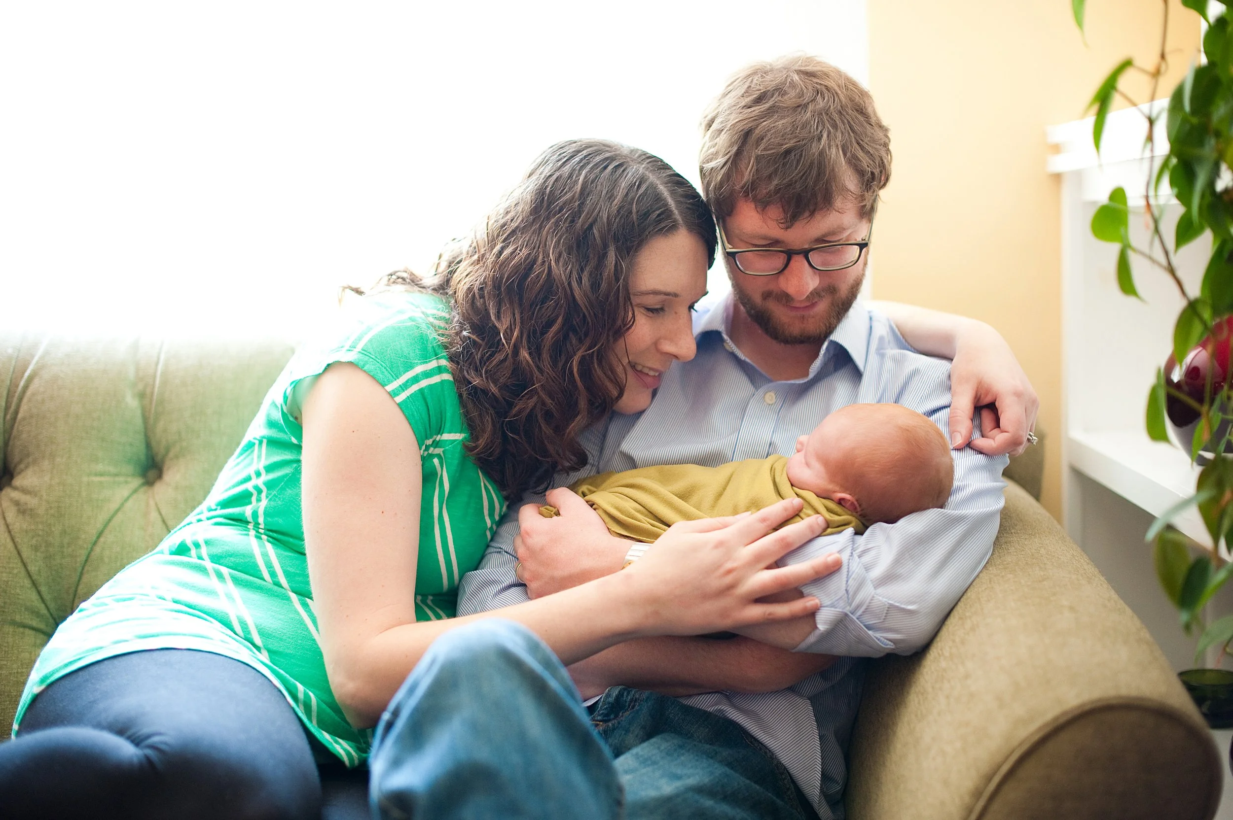 A couple sitting on a beige couch, holding and looking lovingly at a newborn baby wrapped in a yellow blanket.
