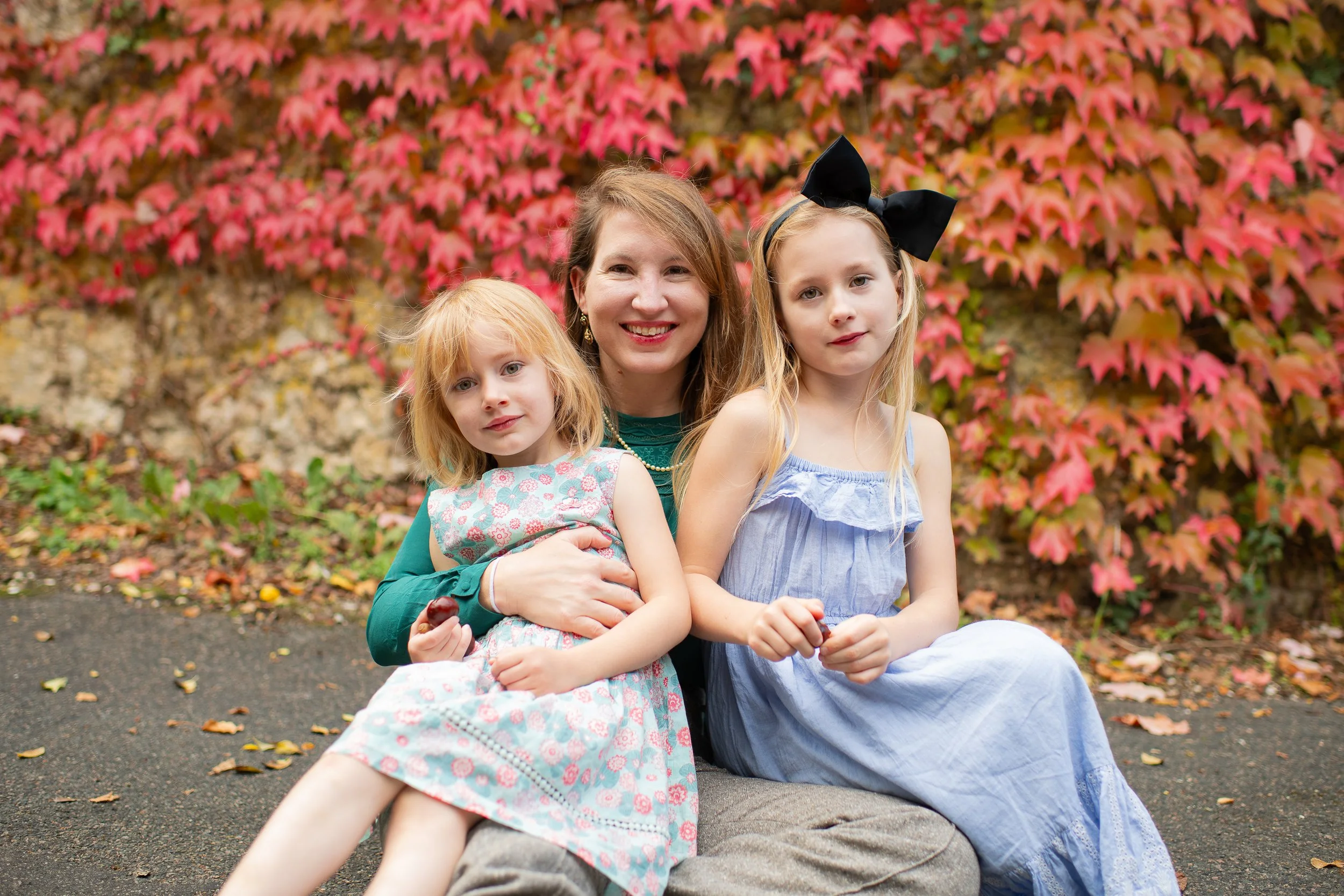 A woman with two young girls sitting together outdoors in front of a red and pink autumnal foliage background.