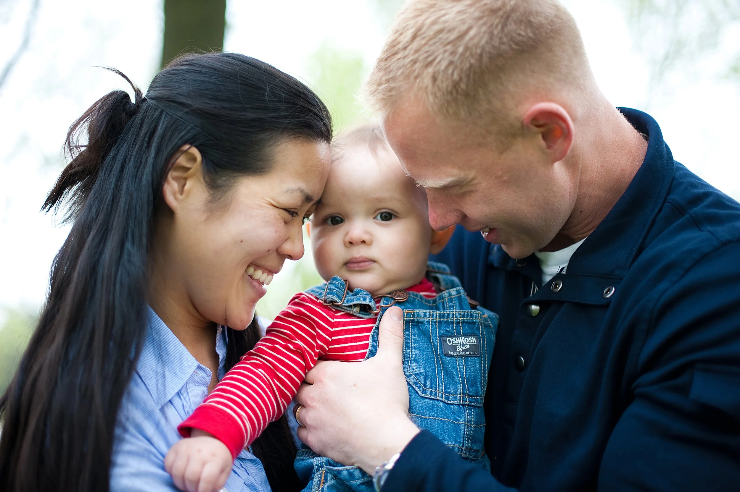 A happy woman and a man smiling at a curious baby girl they are holding between them outdoors.