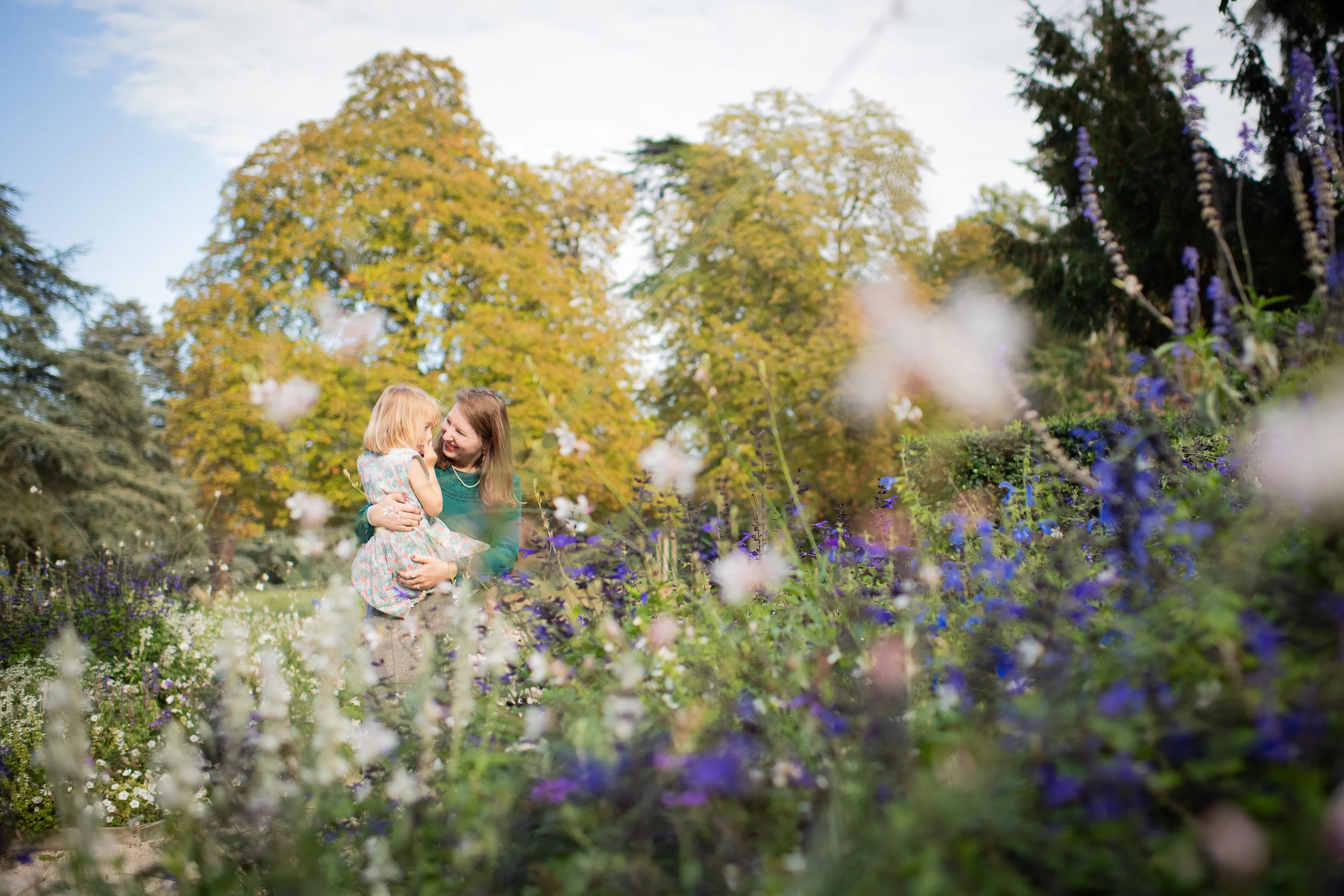A woman and a young girl are outside in a lush garden with colorful flowers and tall trees in the background. The woman holds the girl in her arms, and they are sharing a joyful moment, smiling and touching noses.