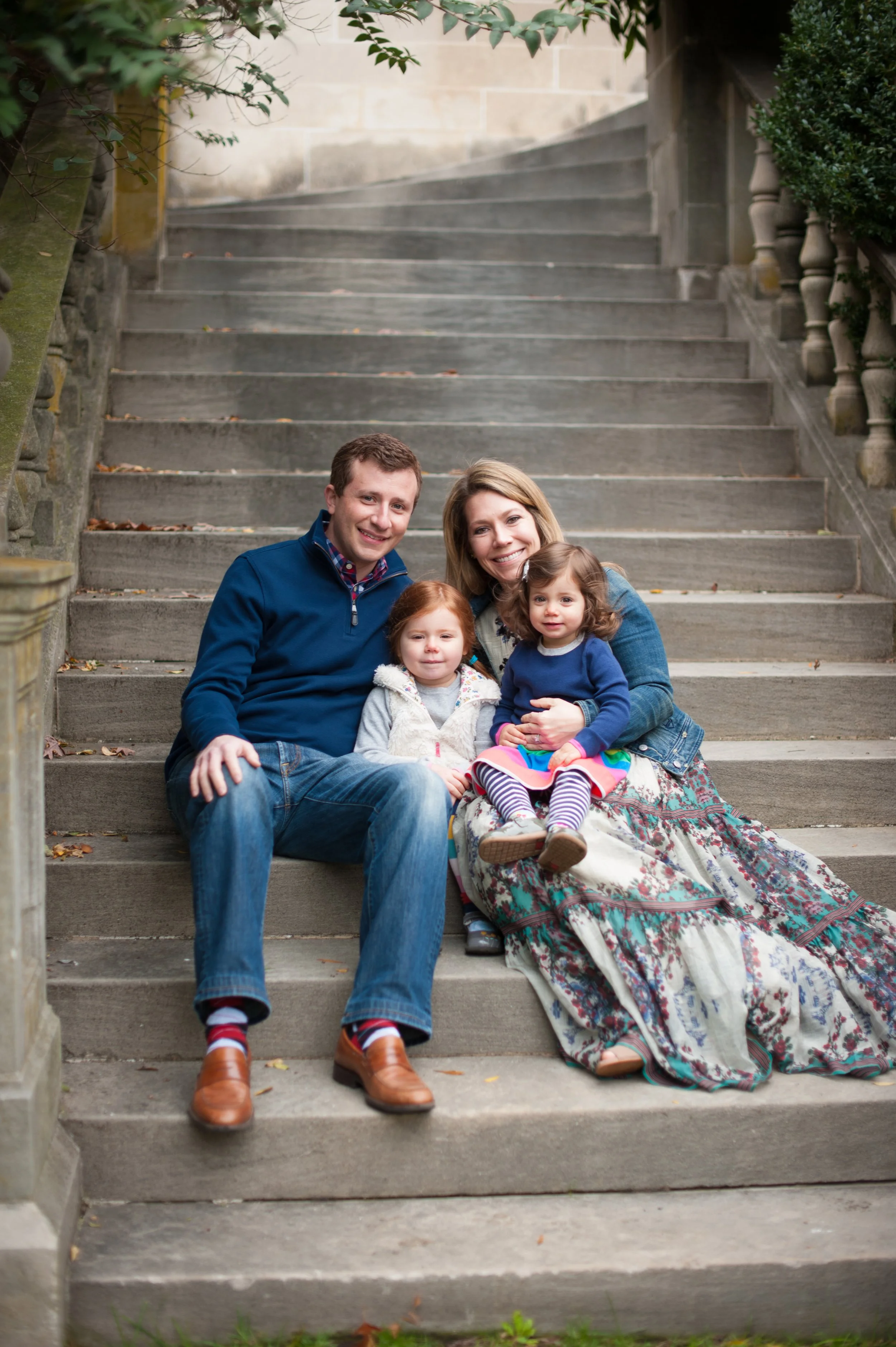 Family of four sitting on outdoor stone steps, smiling at the camera. The father and mother sit with two young daughters between them, all dressed casually with autumn leaves scattered on the steps.