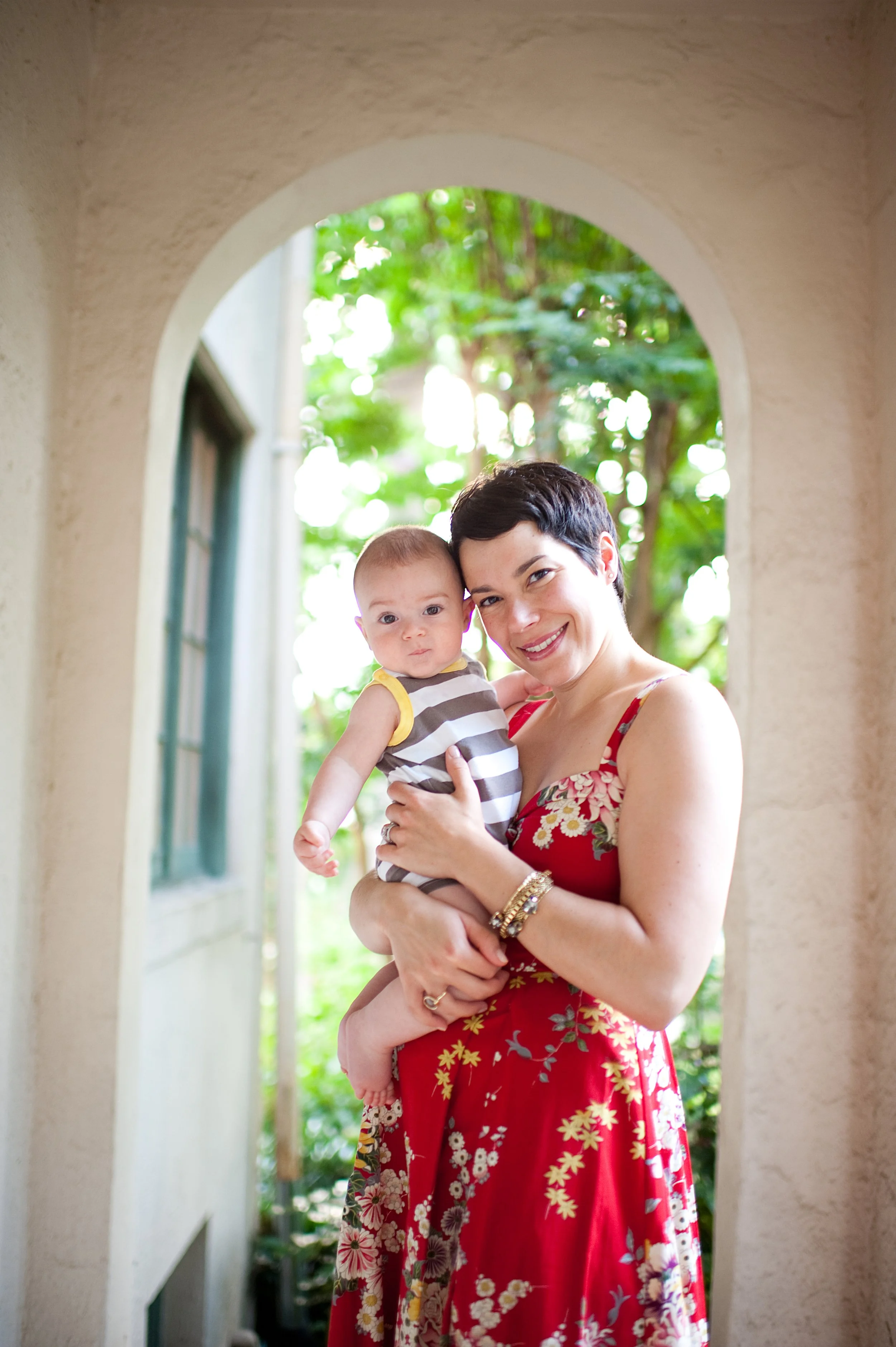 A woman with short dark hair holding a baby with light hair and wearing a striped sleeveless outfit, both smiling, standing in an arched doorway with greenery in the background.
