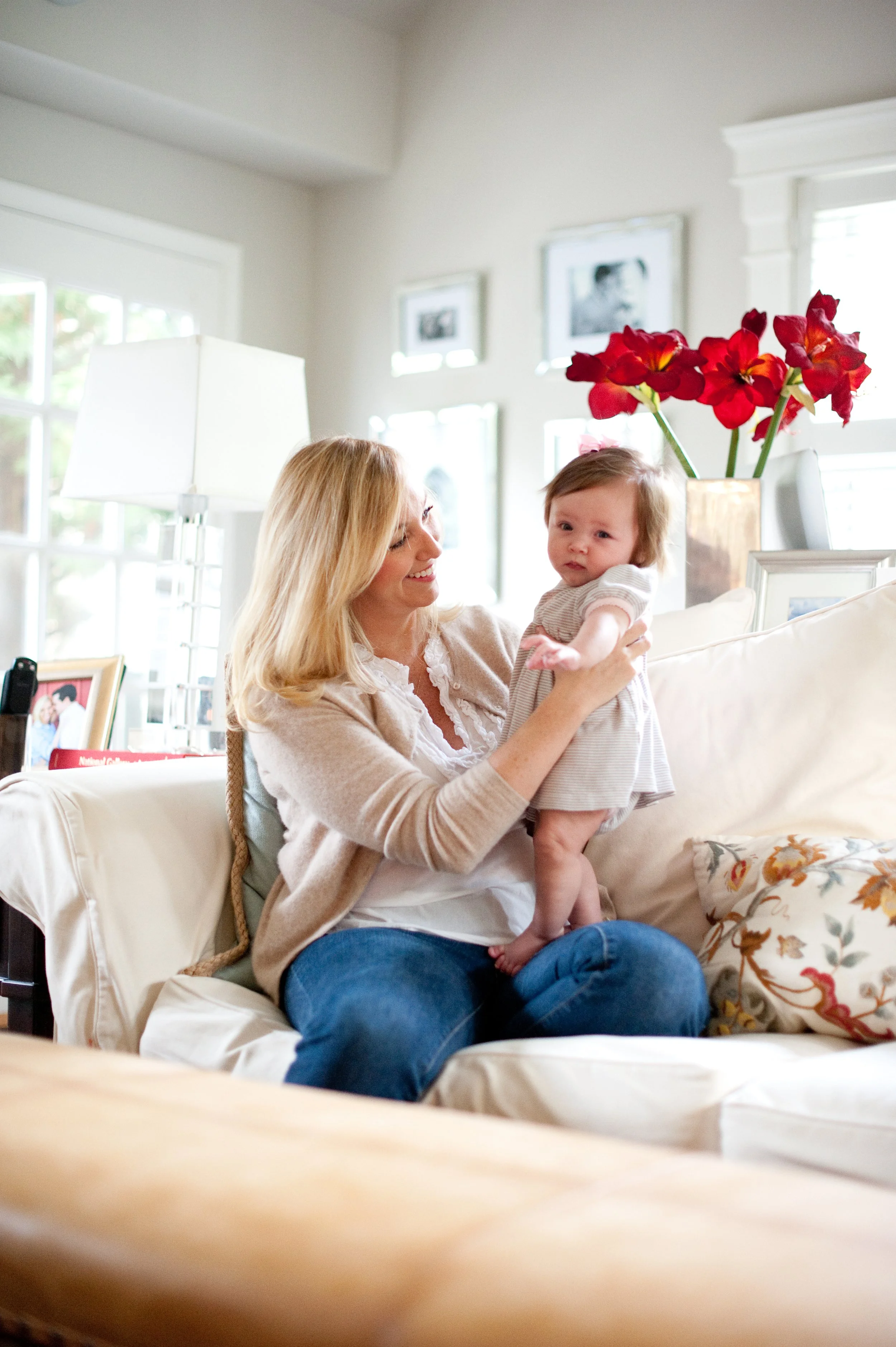 A smiling woman with blonde hair holding a young girl with brown hair on a beige couch in a bright living room with framed photos on the wall and red flowers in a vase.