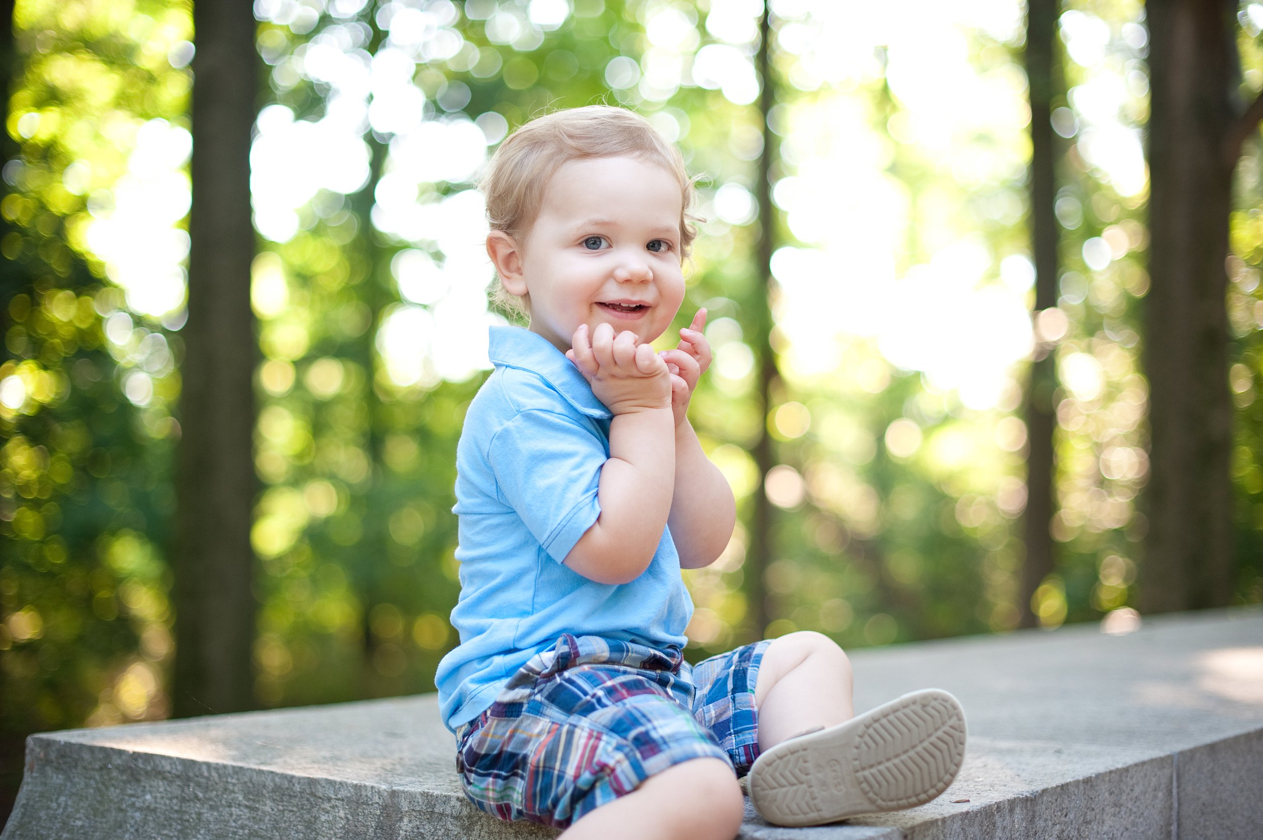 A young boy in a blue polo shirt and plaid shorts sitting on a stone surface outdoors surrounded by green trees, smiling and holding his hands near his face.