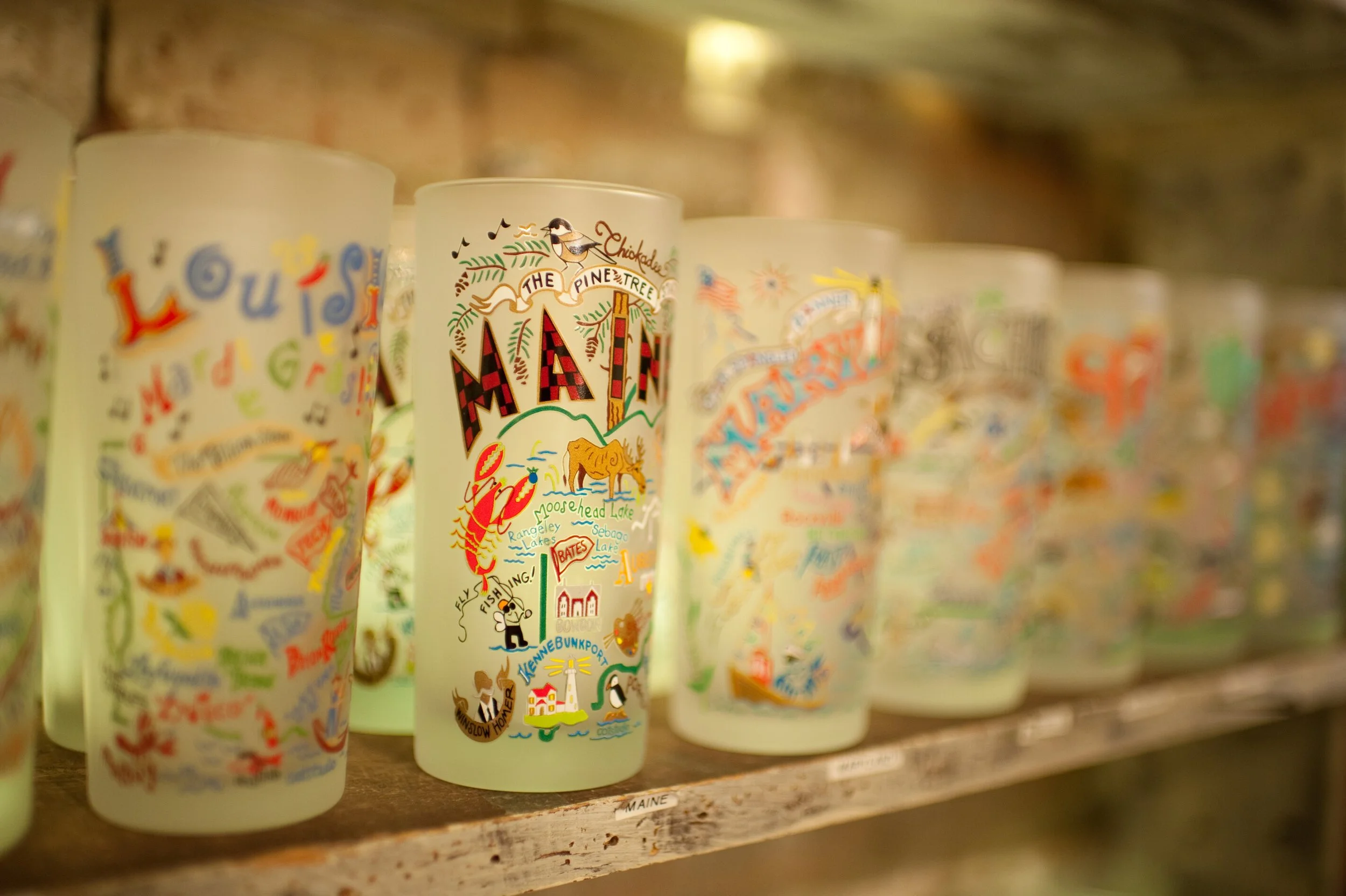 Colorful souvenir glasses with illustrations and text related to Maine, displayed on a wooden shelf.