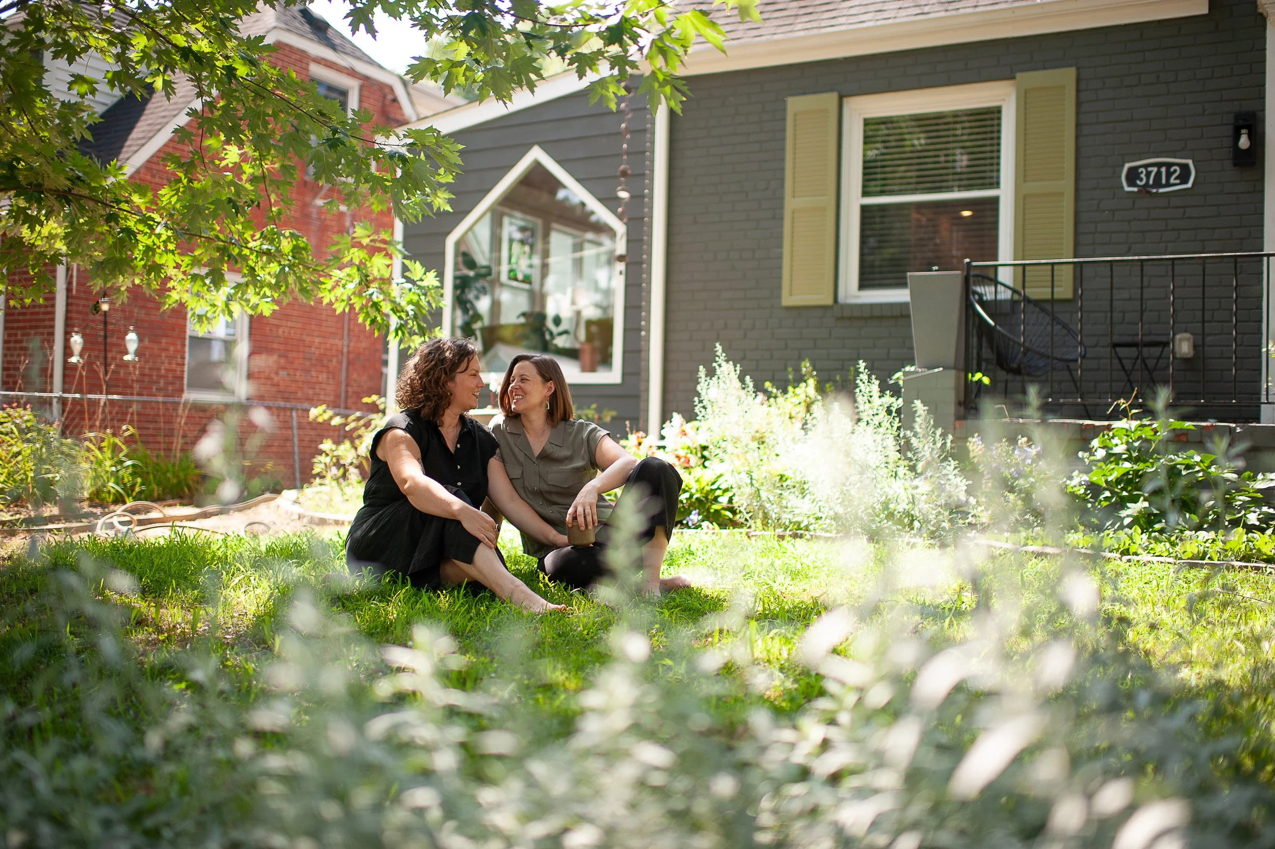 Two women sit together on the grass in a backyard garden, smiling and talking, with a gray house and trees in the background.
