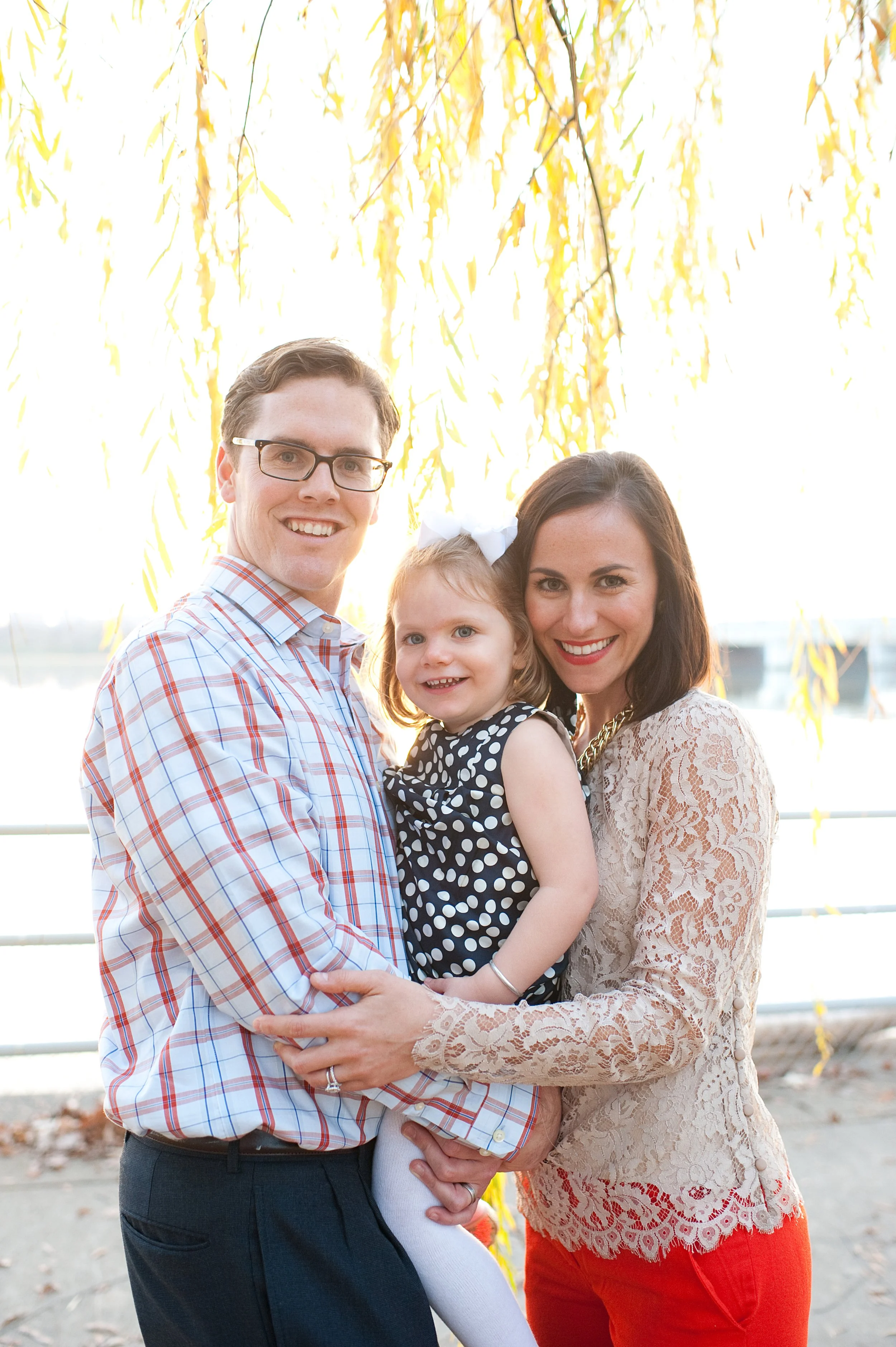 A happy family of three standing outdoors near a railing with autumn leaves behind them, smiling at the camera.