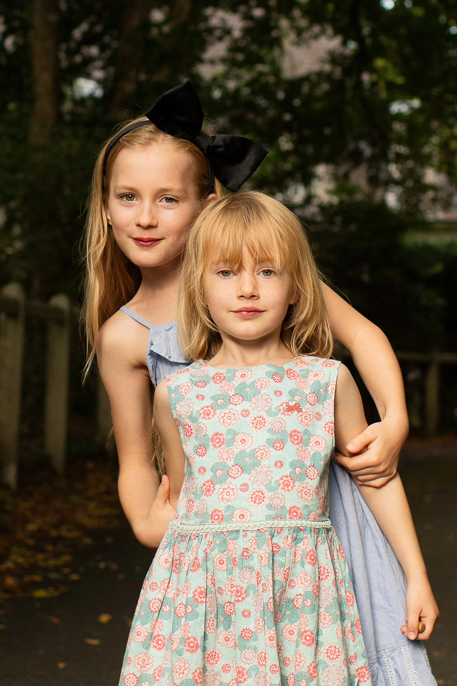 Two young girls standing outdoors in a park-like setting with trees and a pathway. The older girl has red hair, is wearing a light blue dress with a black bow headband, and is smiling. The younger girl has blonde hair, is wearing a floral dress, and 