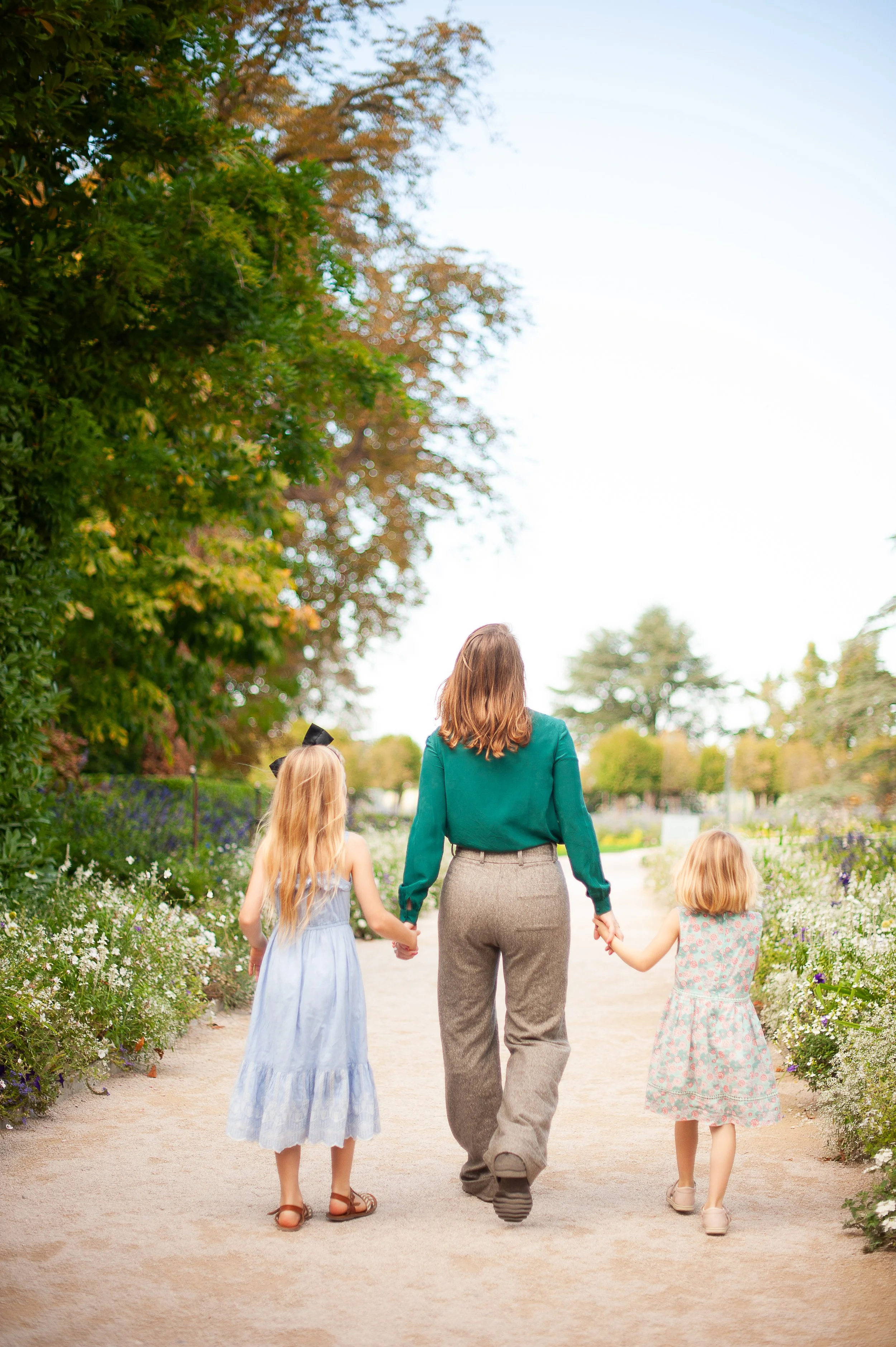 A woman walking with three young girls in a garden filled with white and purple flowers. The woman is holding hands with two of the girls, one girl is wearing a light blue dress and the other a floral dress, while the third girl is walking beside her