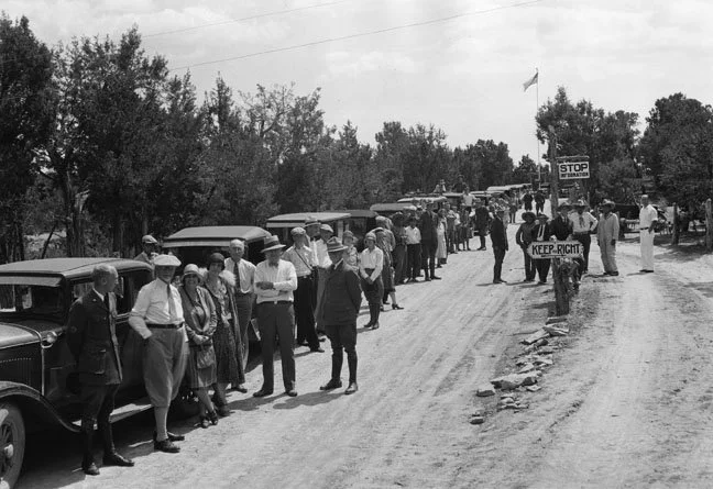 A black and white image of a line of rangers and tourists stand next to cars at Mesa Verde in 1929.
