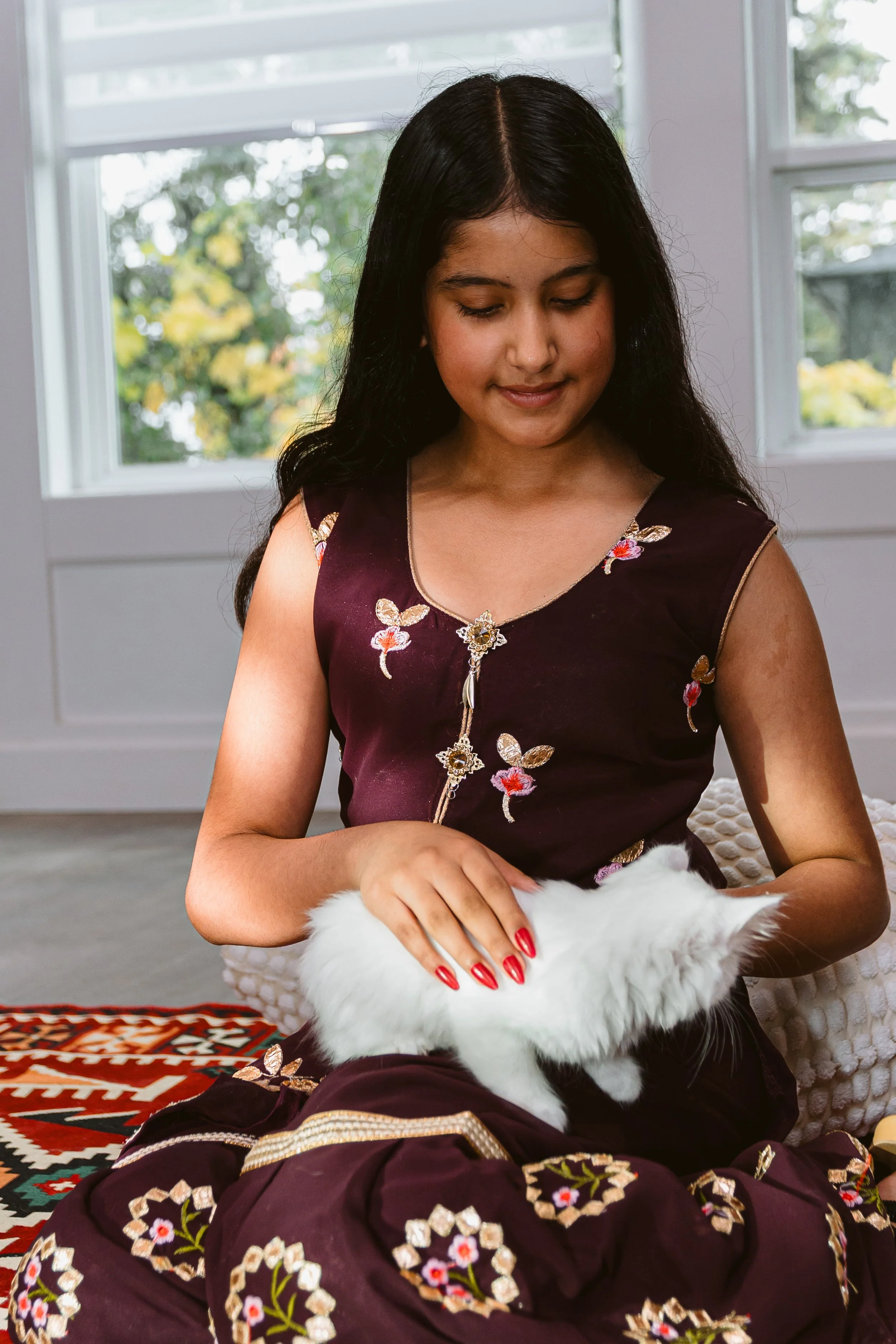 A young girl with long dark hair, wearing a maroon embroidered dress, is sitting and petting a fluffy white cat.