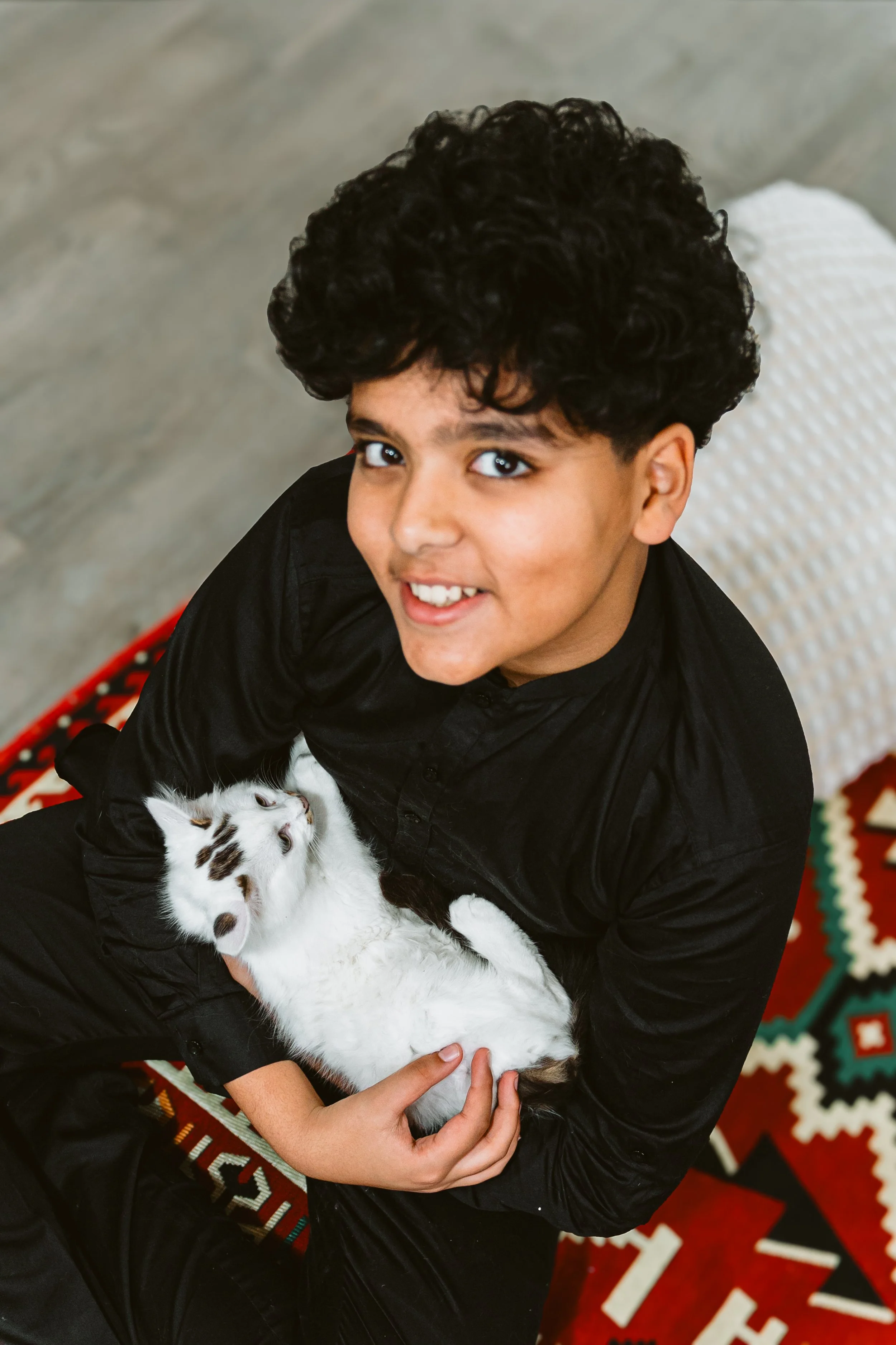 A young boy with curly dark hair holding a white kitten with brown spots. They are sitting on a colorful patterned rug and looking up at the camera.