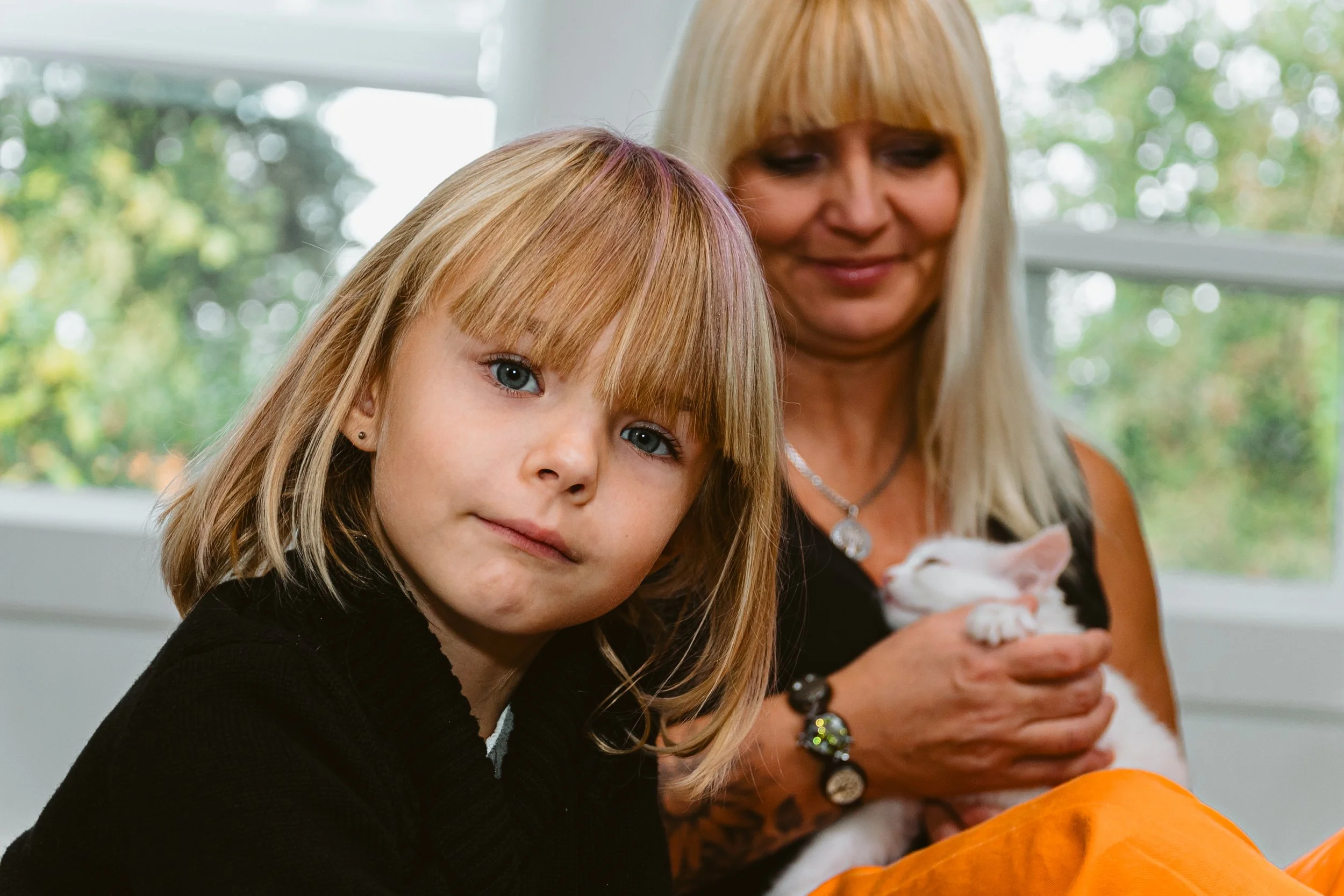 A young girl with blonde hair and blue eyes looks at the camera, sitting beside a woman with blonde hair holding a white kitten, with a window and green foliage in the background.