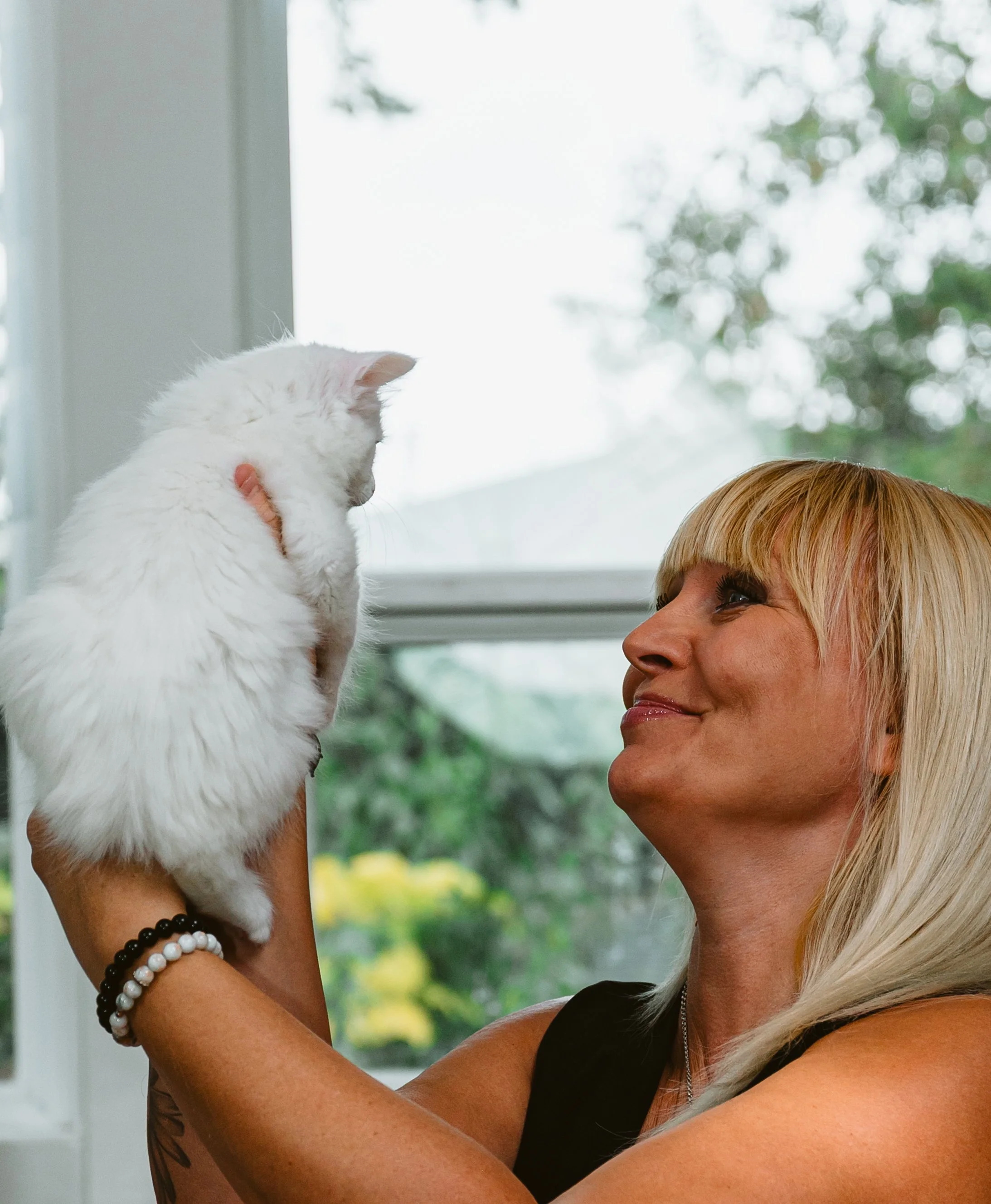 A woman with blonde hair holding a white fluffy cat up close to her face, indoors near a window with trees outside.