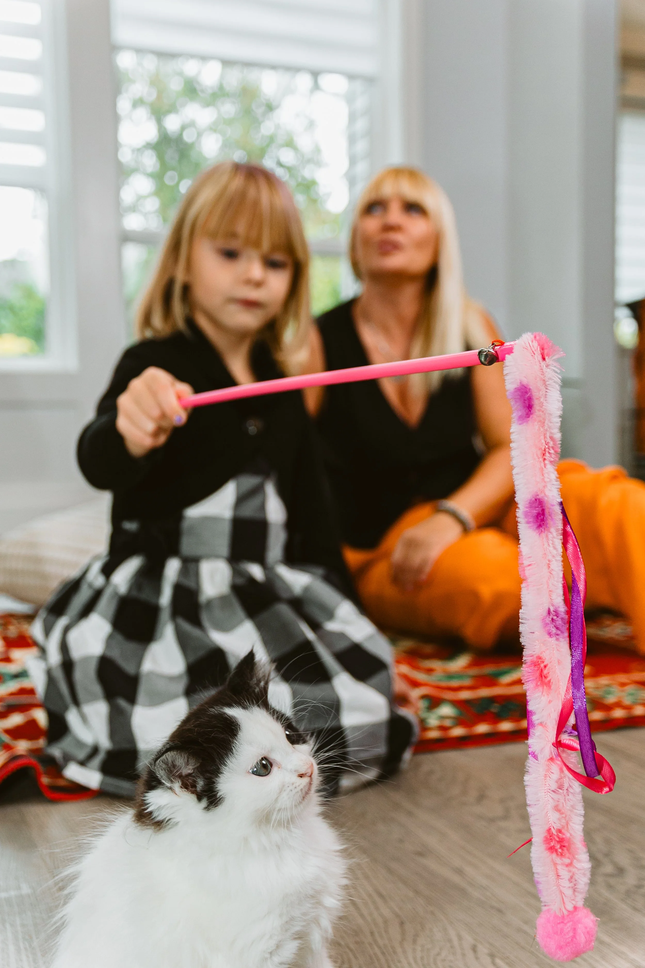 A young girl and an older woman sitting on a colorful rug indoors, with a black and white cat in the foreground. The girl is holding a pink toy with a fluffy attachment, possibly a cat toy, and appears to be playing with the cat.