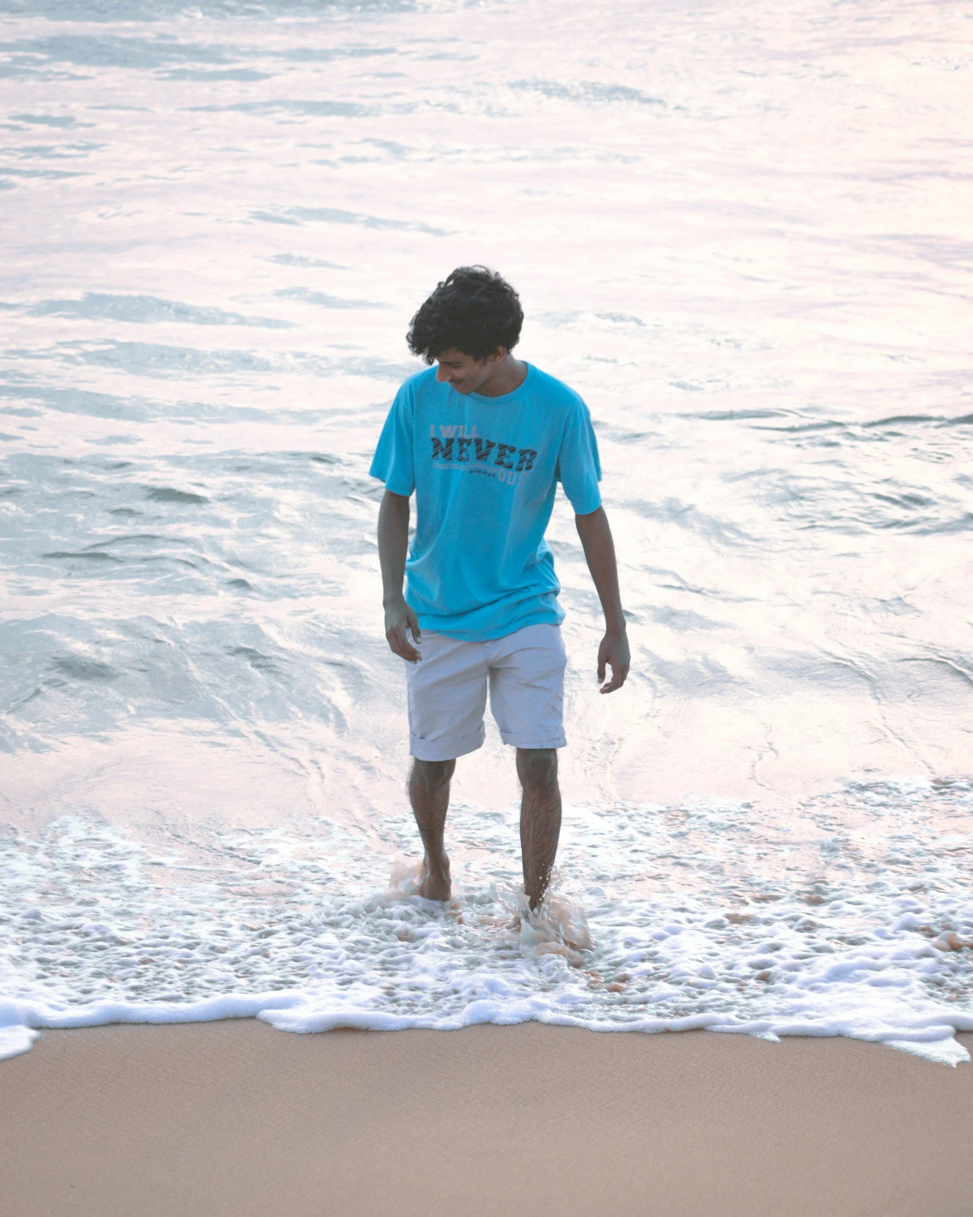 A teen boy in a blue T-shirt and white shorts walking along the water at a Florida beach during sunset. He feels peaceful, calm, and confident. Online stuttering therapy has helped him to speak smoother and be braver in his communication.