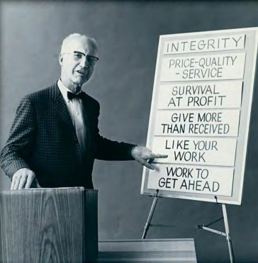 Man in a suit pointing to a whiteboard with business principles listed, including integrity, price-quality-service, survival at profit, give more than received, like your work, work to get ahead.